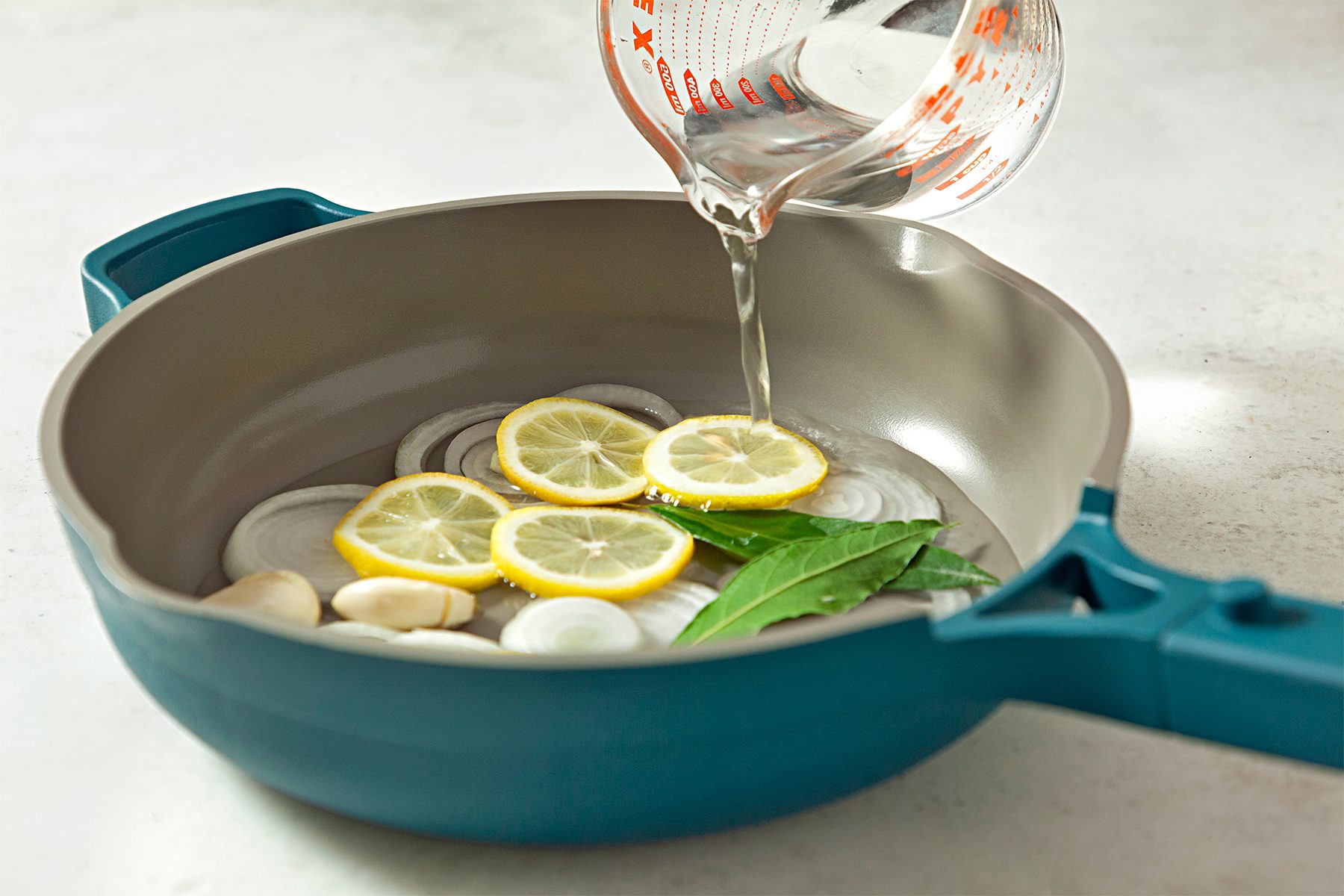 A turquoise frying pan with a non-stick interior contains sliced onions, lemon slices, garlic cloves, and bay leaves. A hand is pouring liquid from a measuring cup into the pan, 