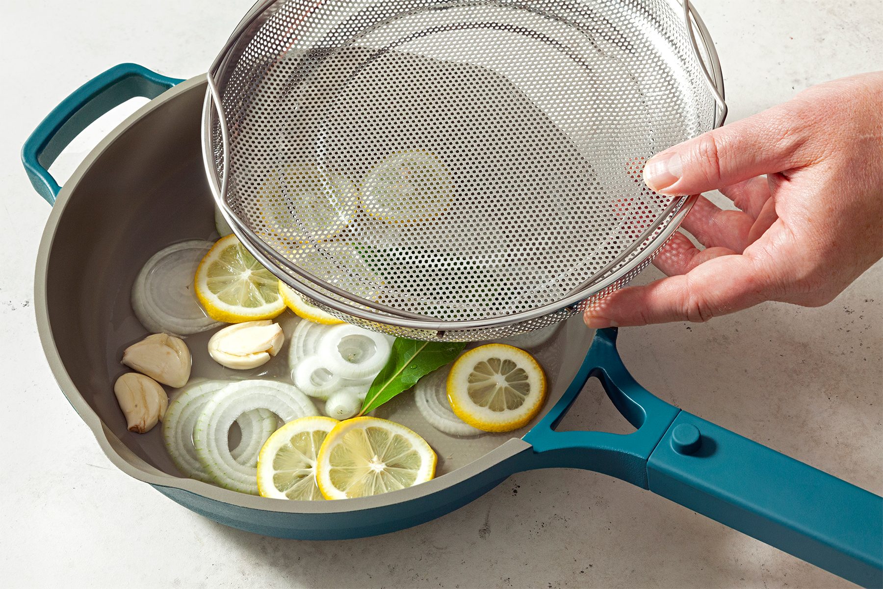 A person holding a mesh lid above a turquoise frying pan containing sliced onions, garlic cloves, lemon slices, and a bay leaf.