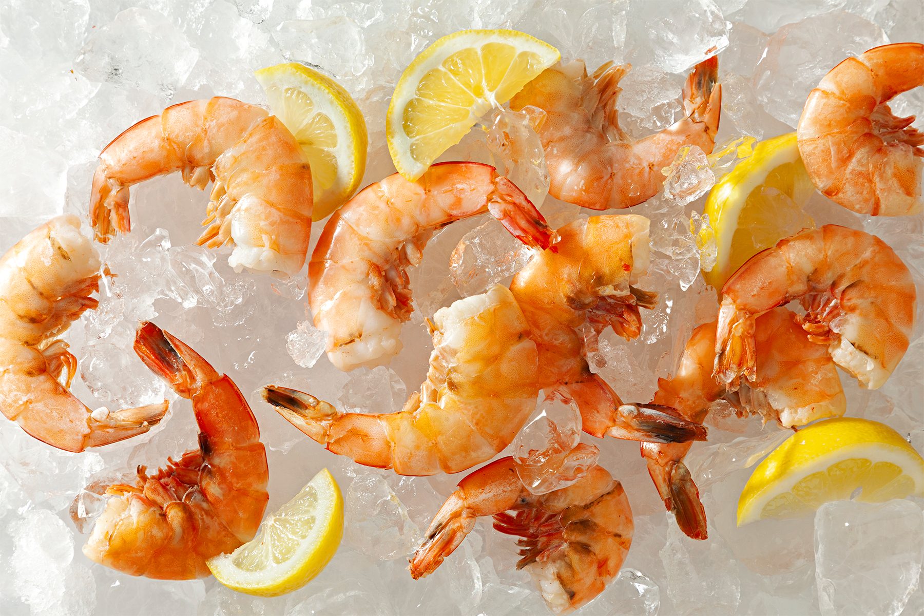 Peeled shrimp with tails on are arranged on a bed of crushed ice, accompanied by lemon wedges. The scene is brightly lit, showcasing the fresh, vibrant colors of the shrimp and lemons.