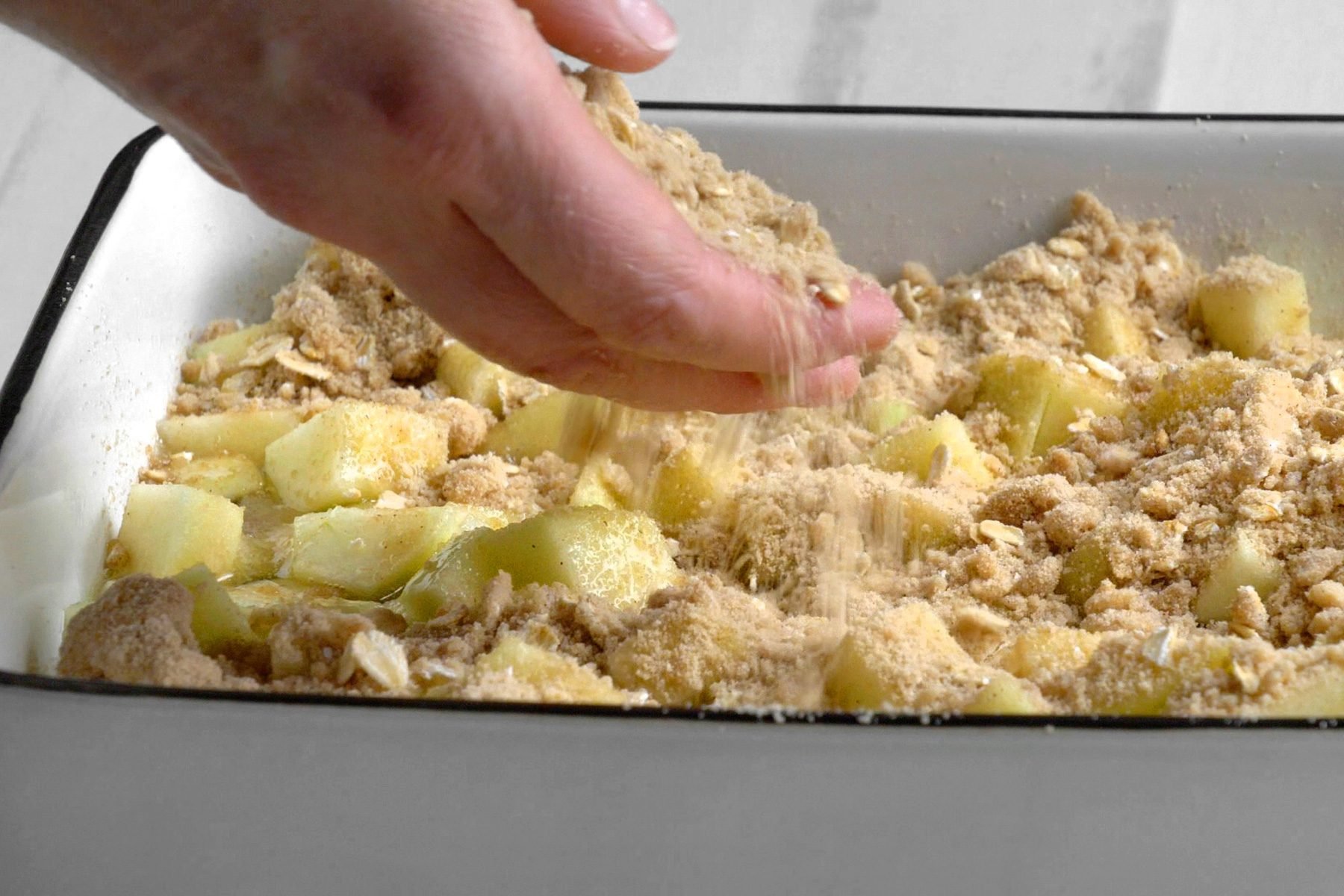 A hand sprinkles the crumb mixture over the layer of chopped apples in a baking dish.