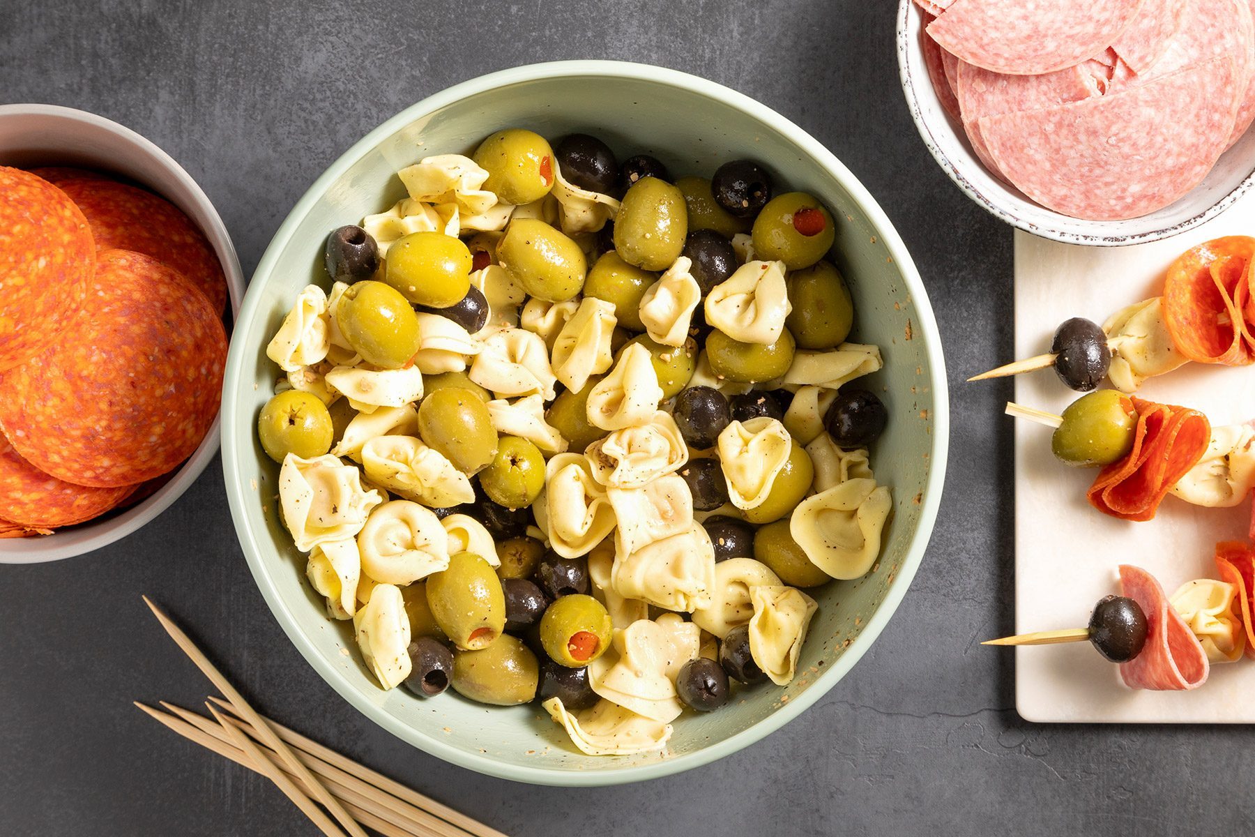 overhead shot; grey textured background; in a large bowl, combine the tortellini, olives with short skewer, pepperoni slices and salami pieces in small bowl