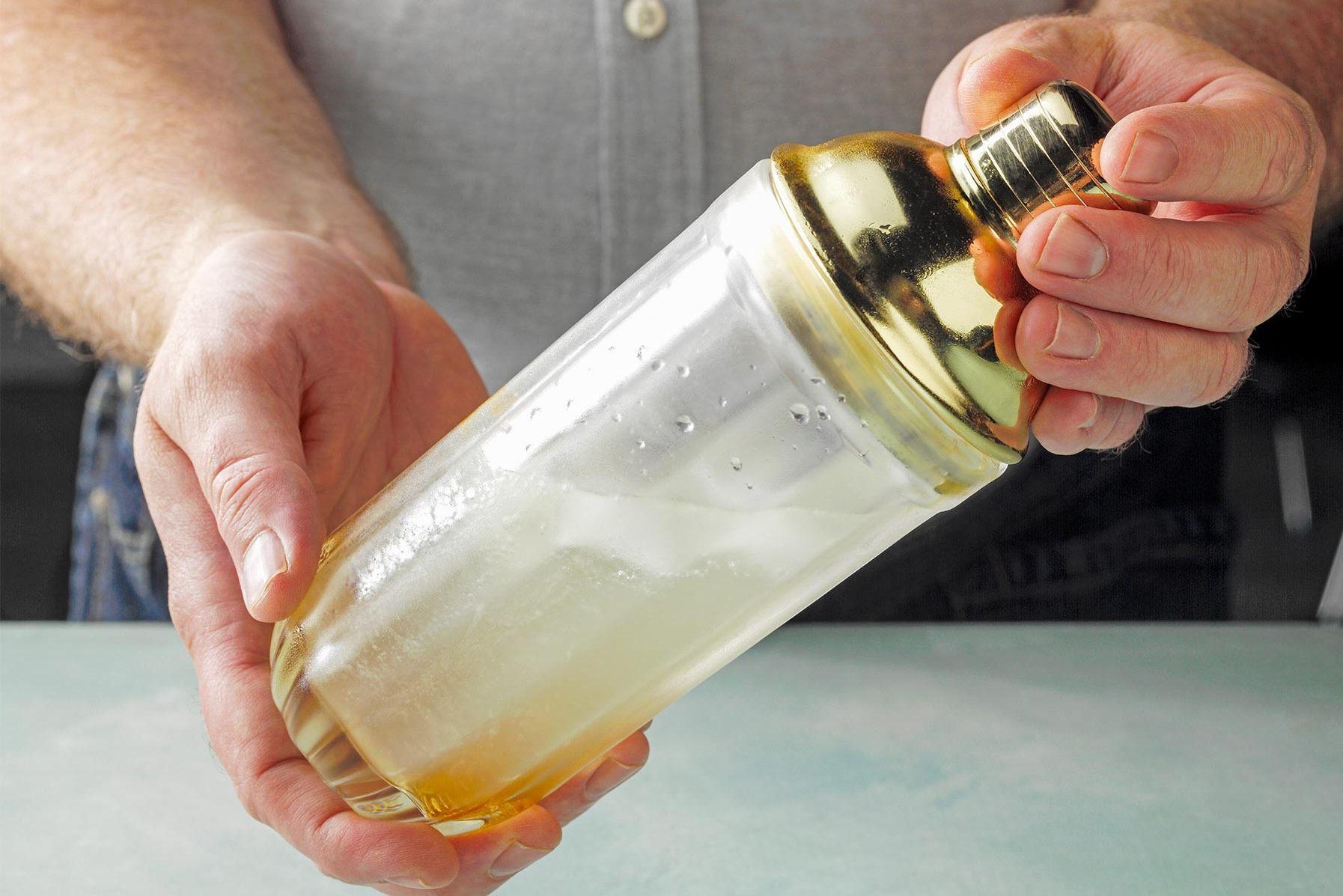 Close-up of a person holding a cocktail shaker. The shaker has a golden top and a transparent body filled with a light-colored liquid and ice.