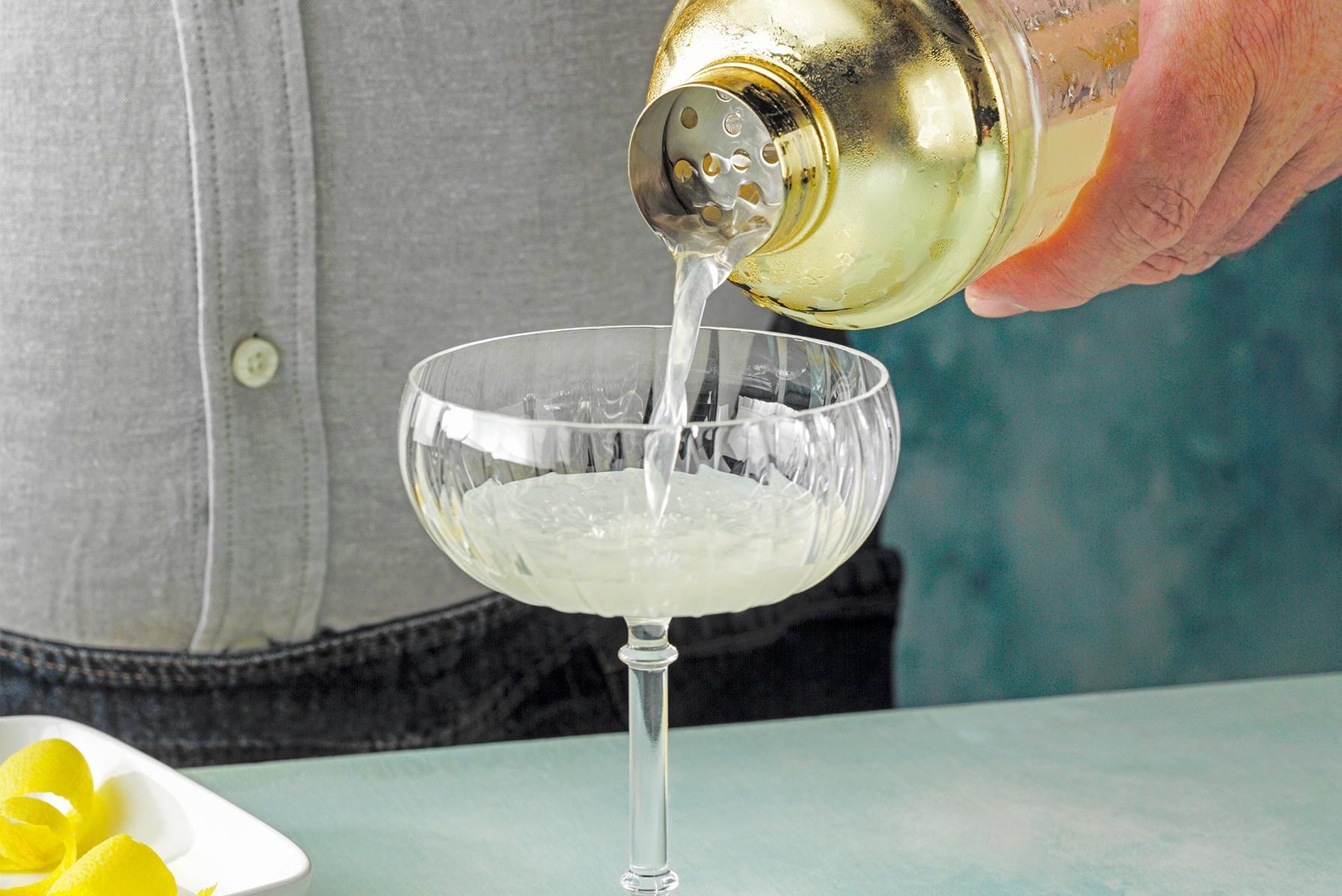 A person is pouring a clear liquid from a gold cocktail shaker into a coupe glass on a light green surface.