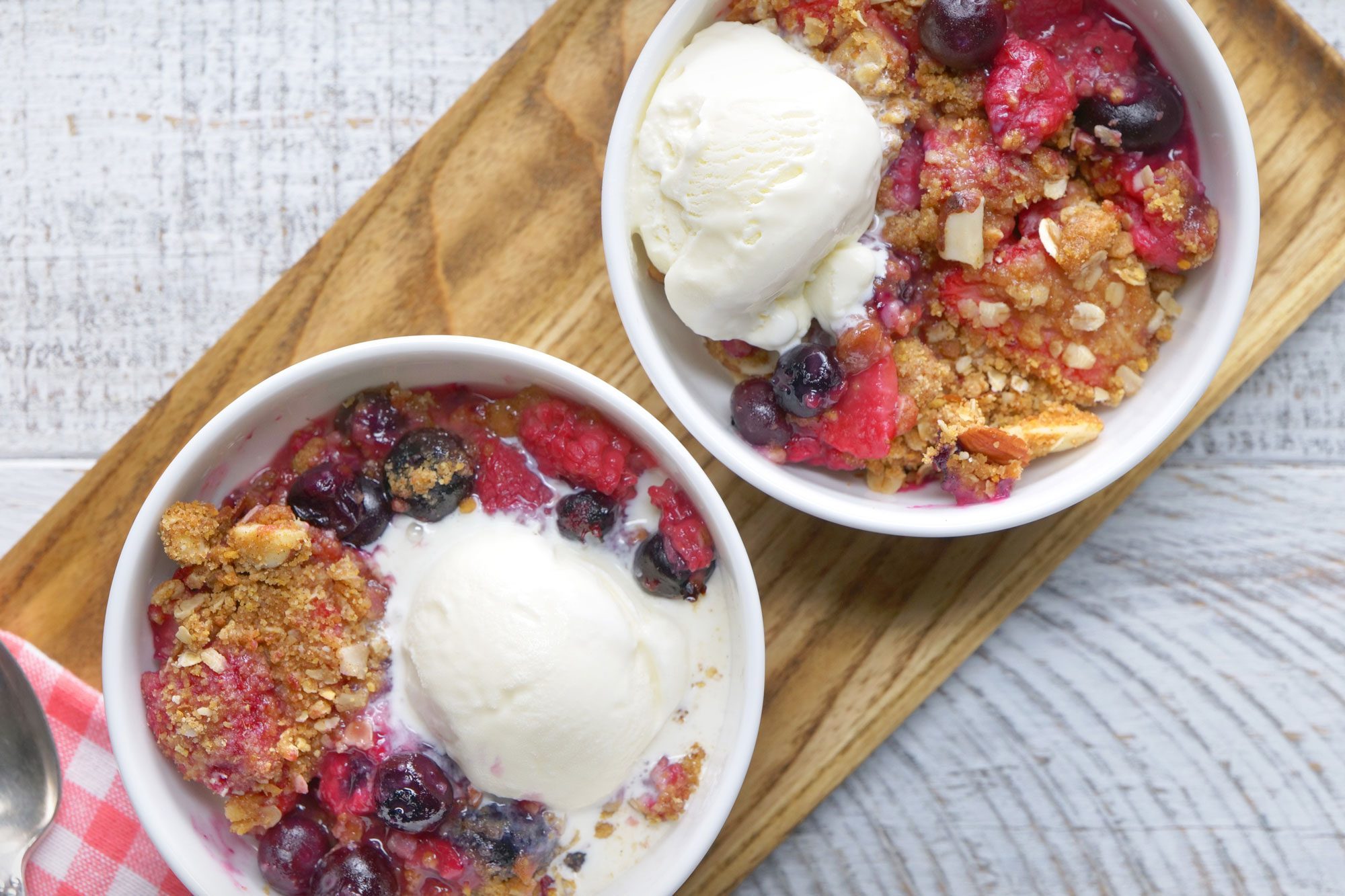 overhead shot; white textured background; 2 bowls of berry crisp garnished with vanilla ice cream on top over wooden board with kitchen towel;