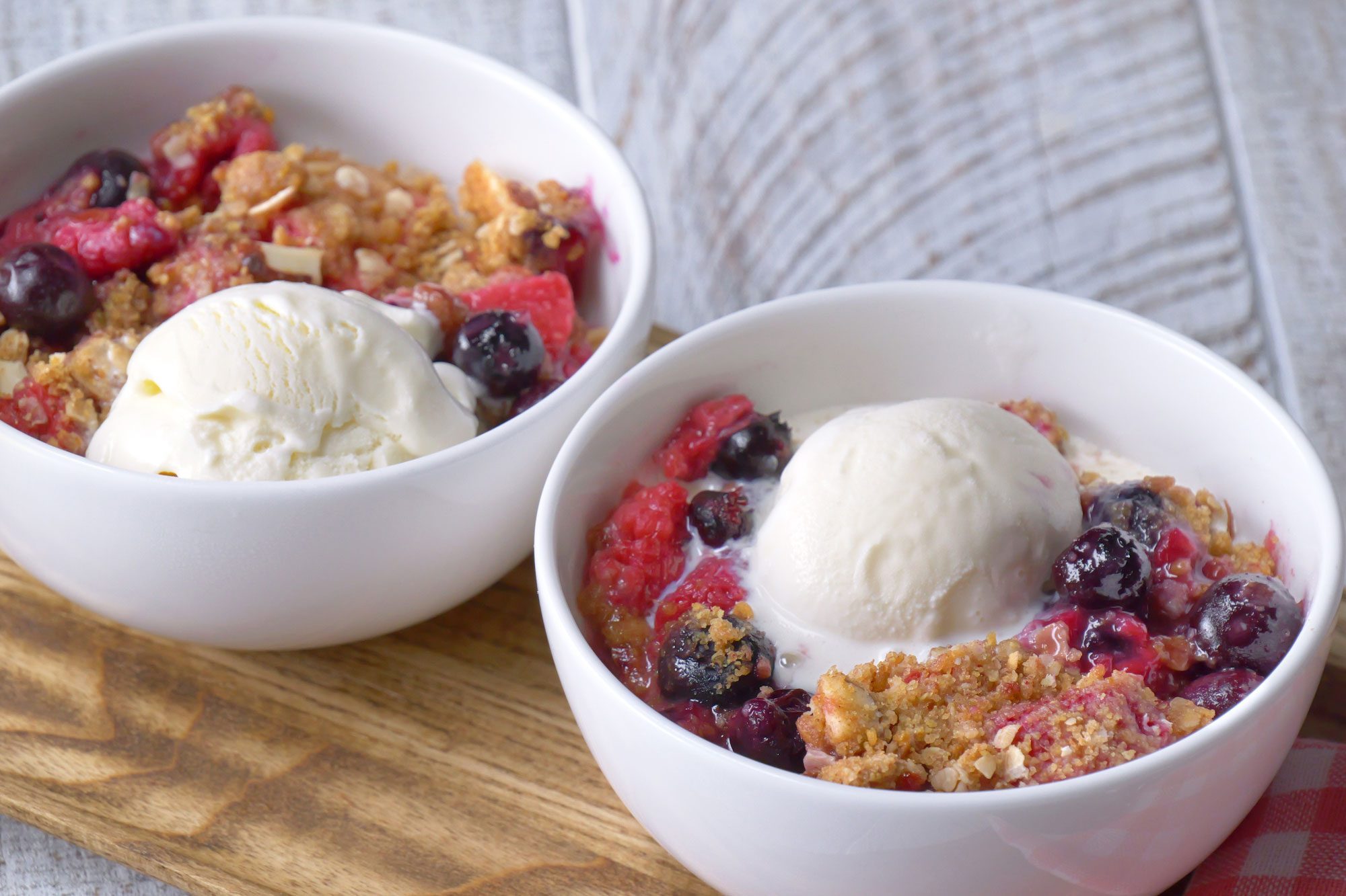 3/4th shot; white textured background; 2 bowls of berry crisp garnished with vanilla ice cream on top over wooden board with kitchen towel;