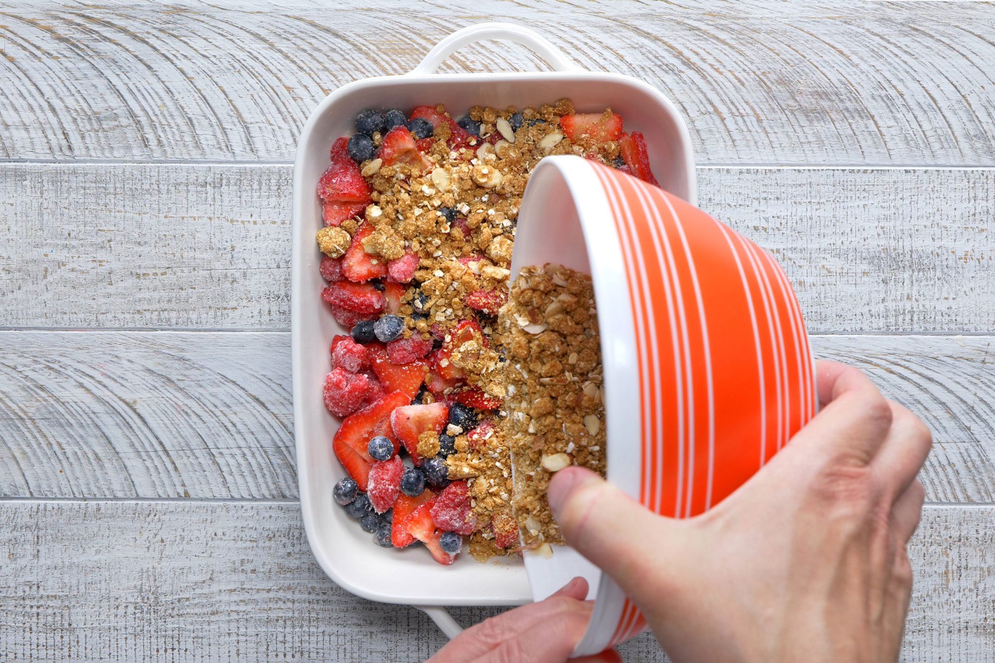 overhead shot; white textured background; Sprinkling berry crisp mixture over berries;