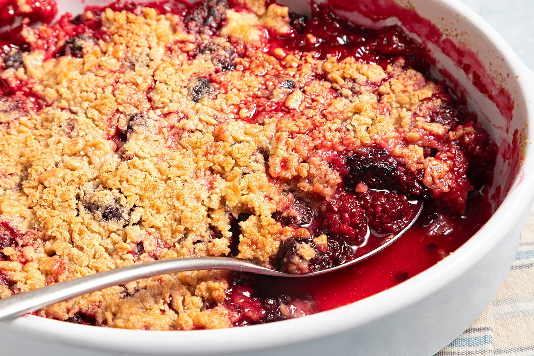Close shot of Blackberry Crumble; in baking dish; serving spoon; kitchen towel; blue marble background