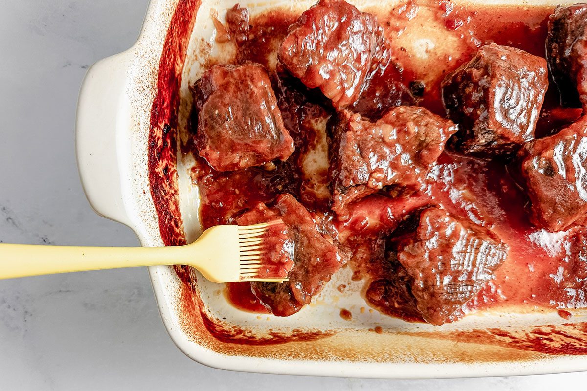 Taste of Home boneless beef short ribs brushed with sauce in a white ceramic baking dish on a marble surface.