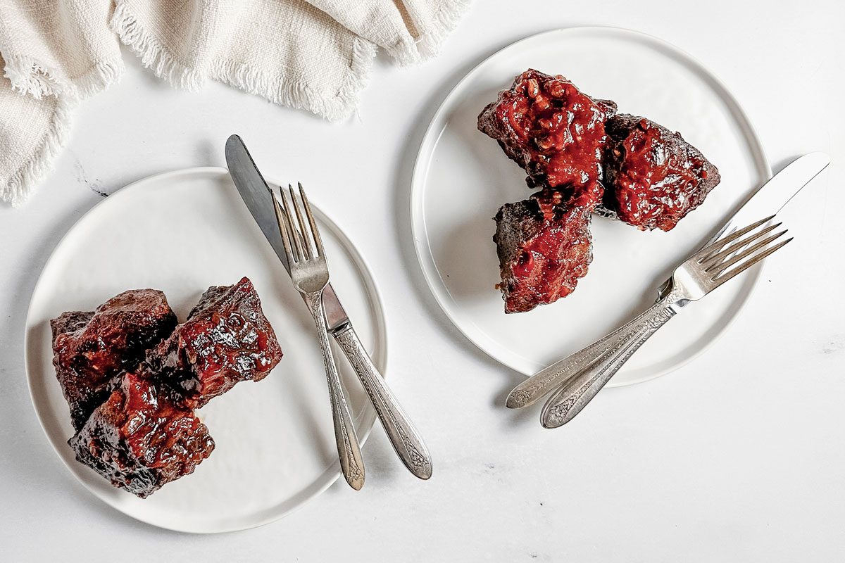 Taste of Home boneless beef short ribs on white ceramic plates with vintage silver cutlery next to a white cloth napkin.