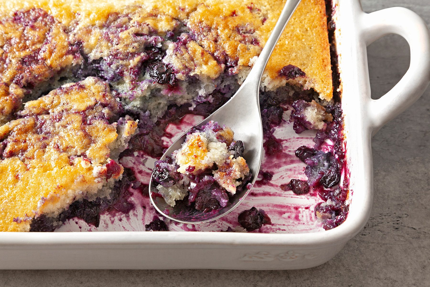 3/4th shot; close shot; grey textured background; Buttery Blueberry Cobbler in a white rectangular baking dish with silver spoon;