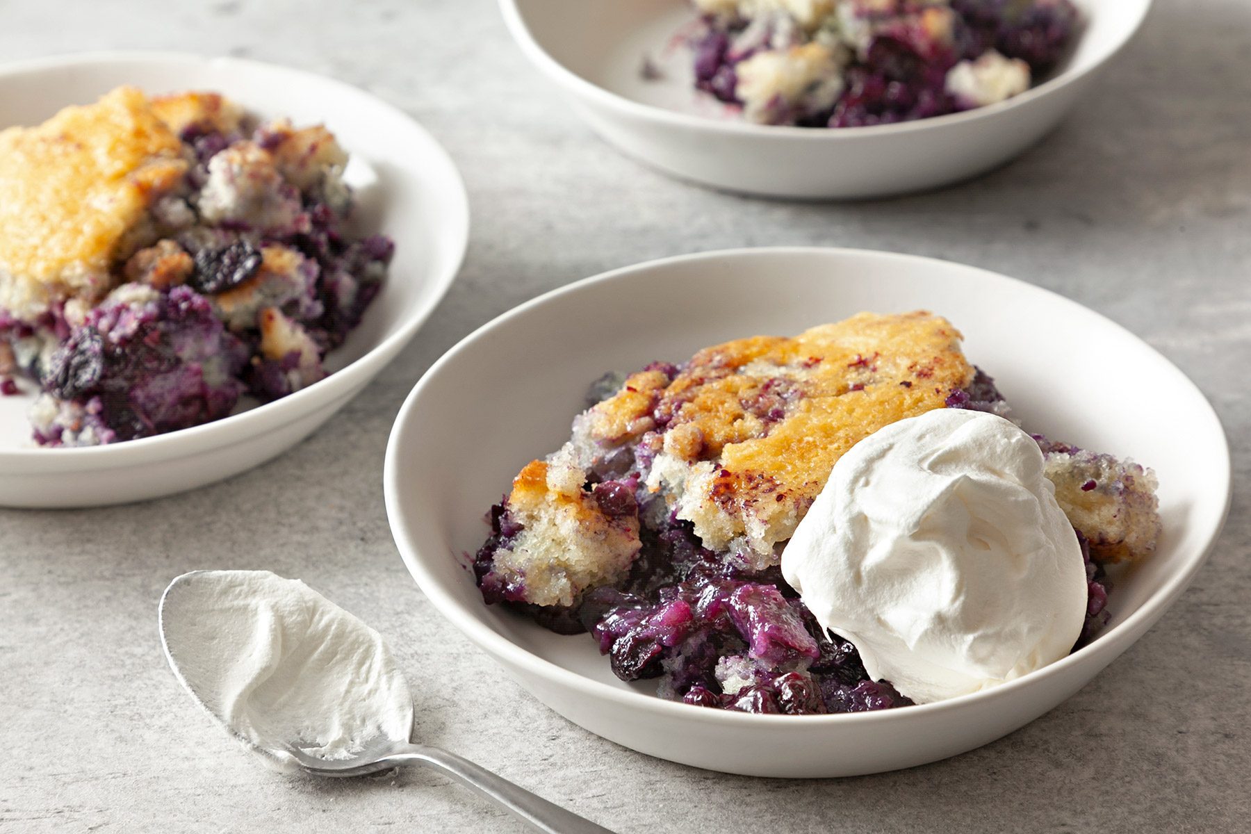 3/4th shot; grey textured background; Buttery Blueberry Cobbler in 3 small bowls served with whipped cream with silver spoon;