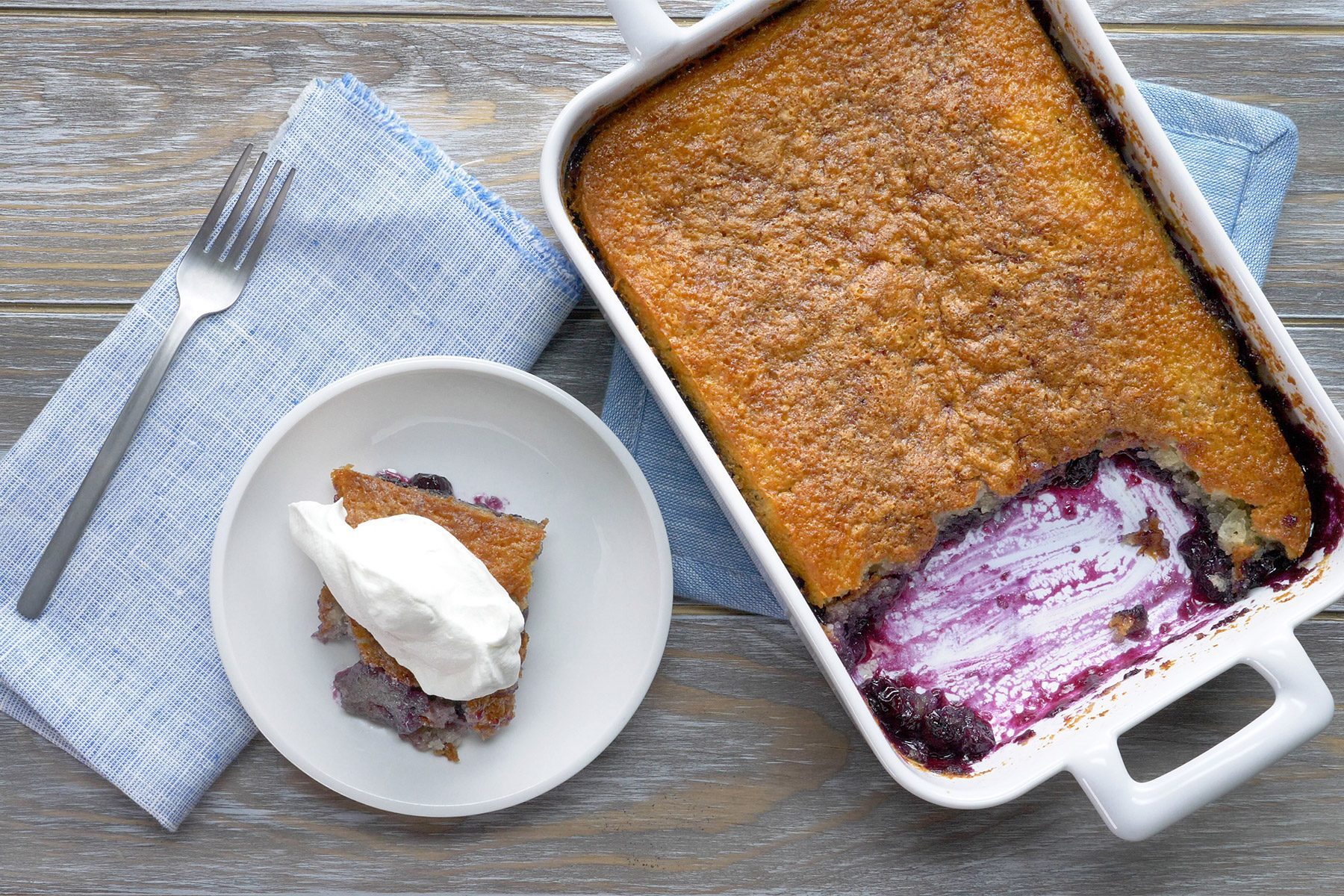 overhead shot; wooden background; baked blueberry cobbler in baking dish; Blueberry Cobbler served in a small white plate with whipped cream; kitchen towel and one silver fork;