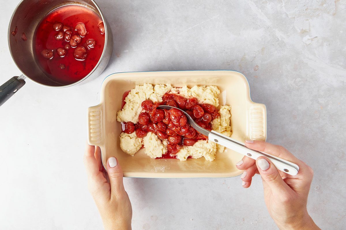 Hands adding cherries to the dough in a baking dish for Cherry Cobbler