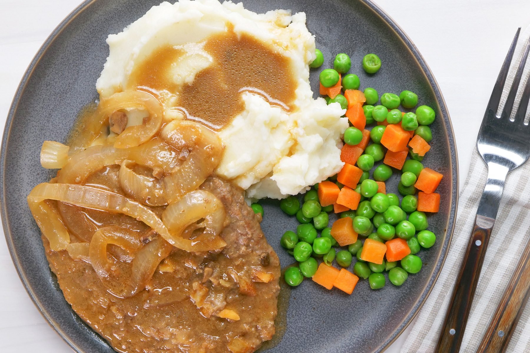 A plate of cube steaks with gravy served over mashed potatoes along with boiled peas and carrot cubes
