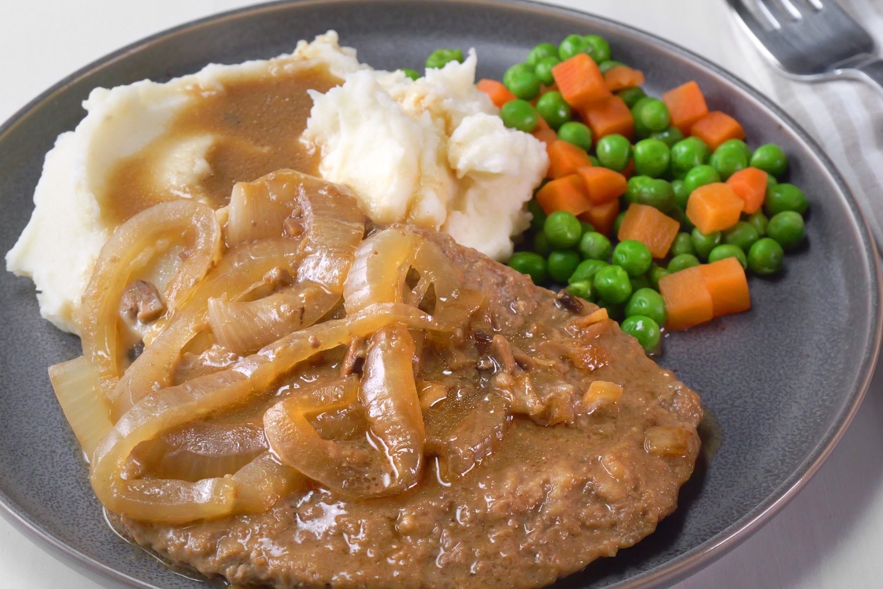 A plate of cube steaks with gravy served over mashed potatoes along with boiled peas and carrot cubes