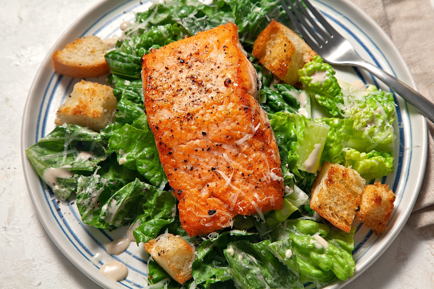 Close shot of Easy Salmon Caesar Salad; served on plates; top each with a salmon fillet; fork; napkin; cream marble background