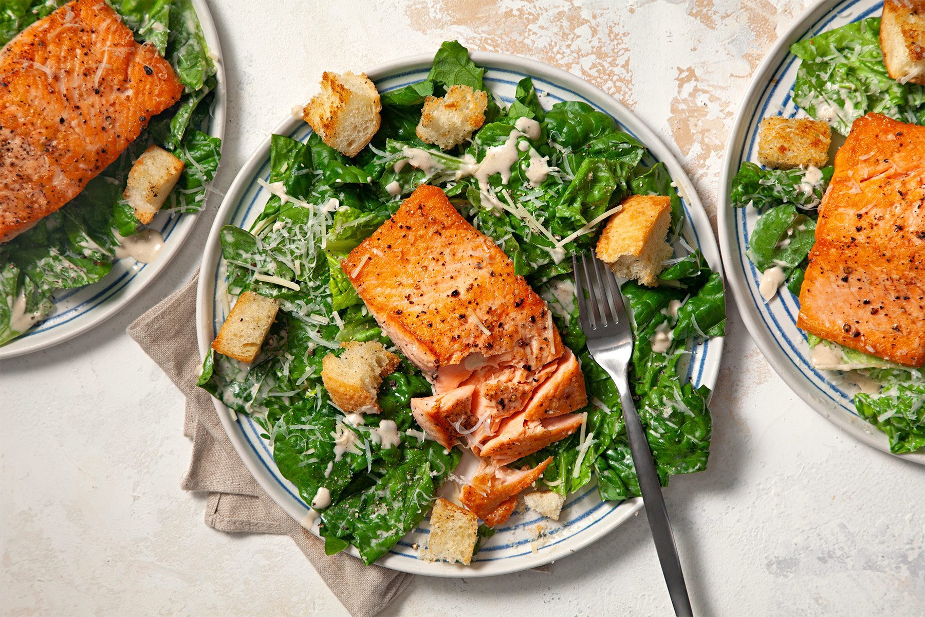 Top view shot of Easy Salmon Caesar Salad; served on plates; top each with a salmon fillet; forks; napkins; cream marble background