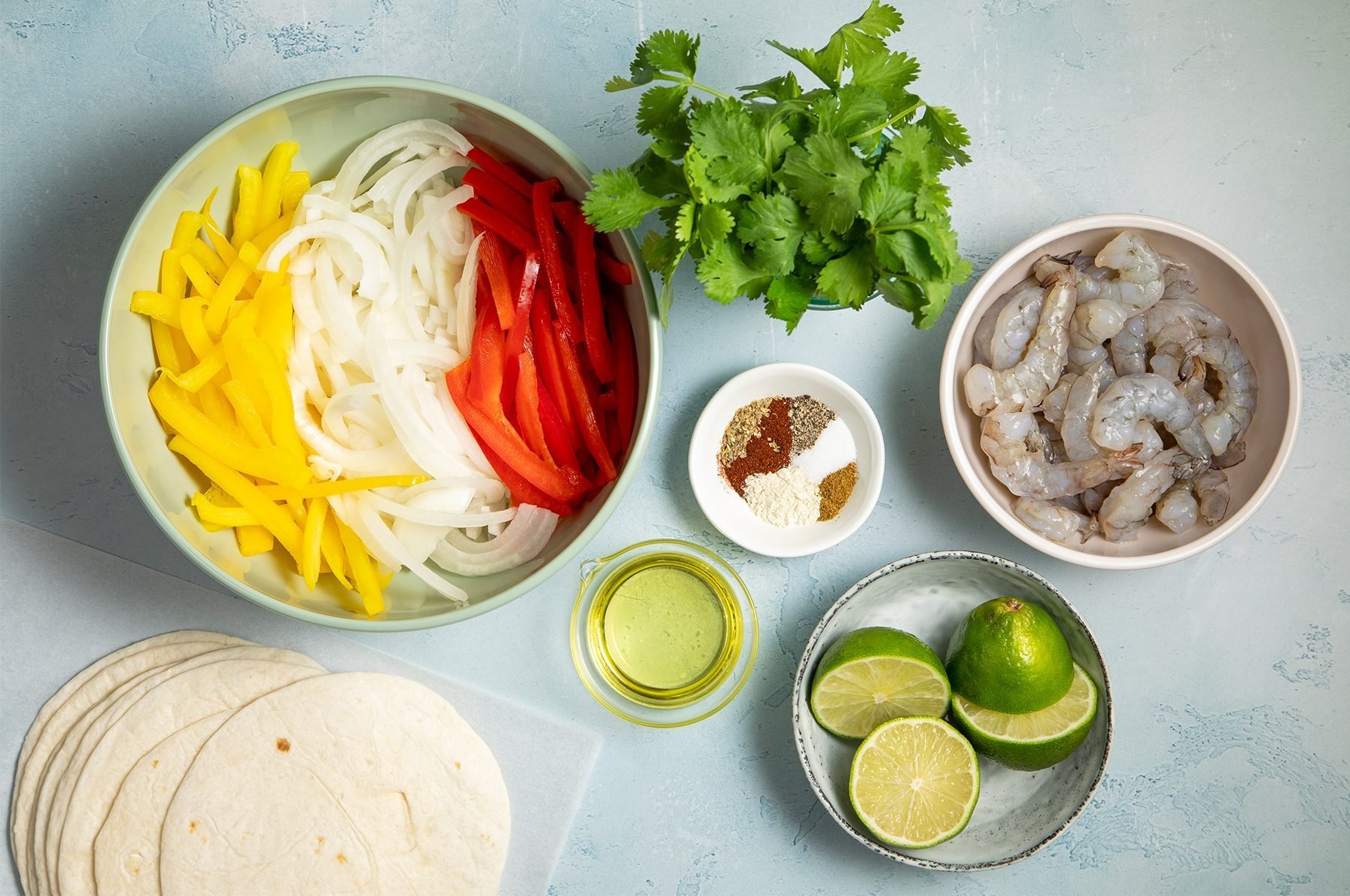A tabletop with ingredients for shrimp tacos, including a bowl of sliced yellow and red bell peppers and white onions, a small bowl of shrimp, a small plate with various spices, two halved limes, fresh cilantro, a small container of oil, and flour tortillas.