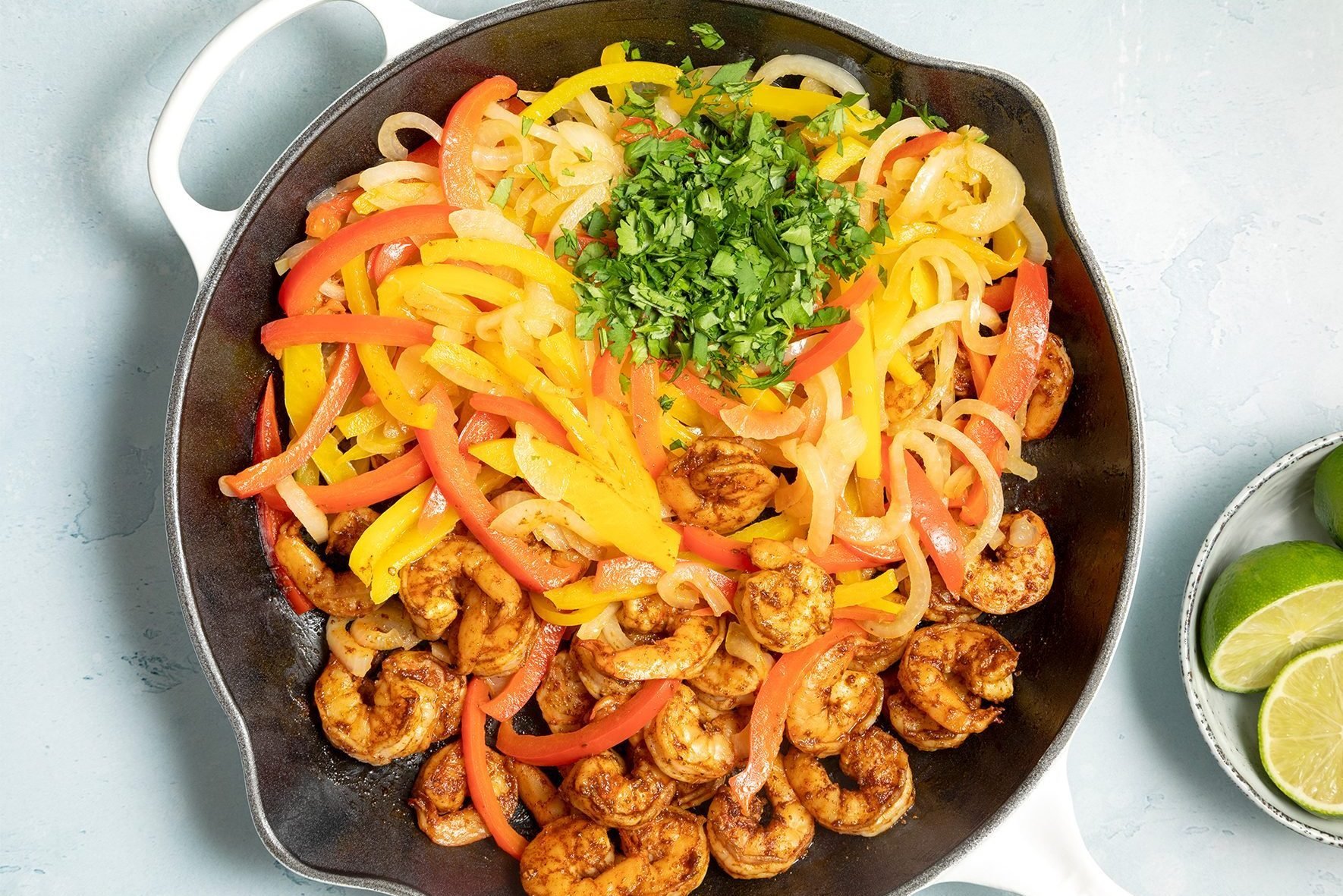 A cast iron skillet filled with seasoned shrimp, sautéed yellow and red bell peppers, and onions. 