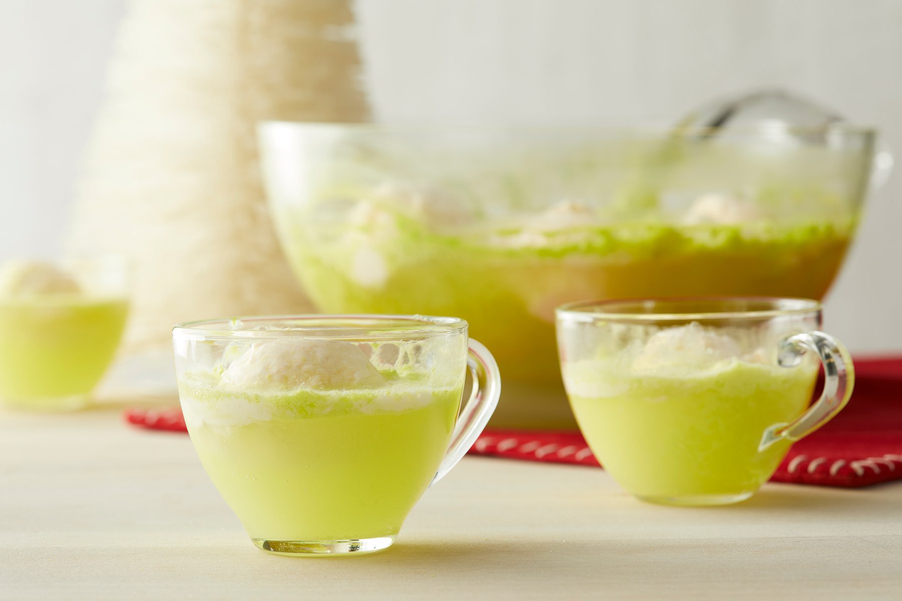 Bright green sherbet punch in clear glass cups with a punch bowl in the background. Each cup contains a scoop of vanilla ice cream floating on top. A red cloth adds a pop of color underneath the punch bowl, with a blurred white object in the background.
