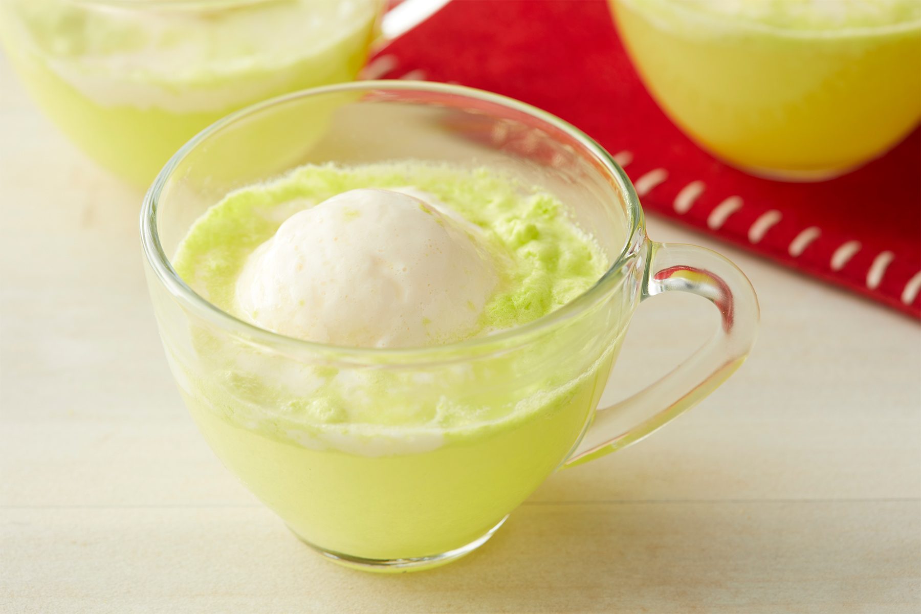A clear glass mug filled with a light green beverage, topped with a scoop of vanilla ice cream. Two more identical mugs are blurred in the background, placed on a red cloth with white stitching. The scene is set on a light-colored surface.