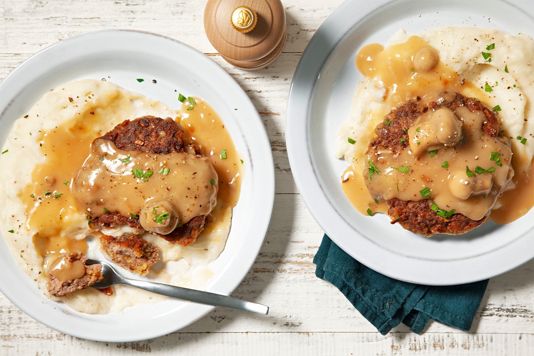 overhead shot; wooden background; Hamburger Steaks with Mushroom Gravy served with mashed potatoes in a white plate with silver spoon over green kitchen towel;