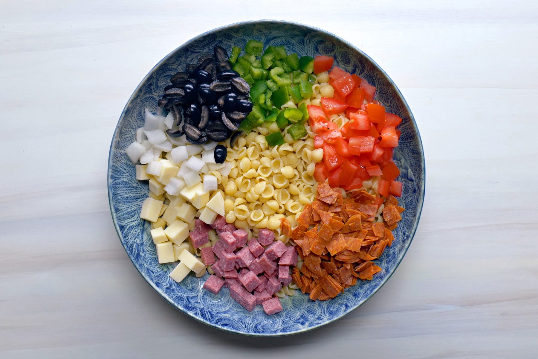 Ingredients including capsicum, pasta, tomatoes, meat, olives for Italian pasta salad in a plate