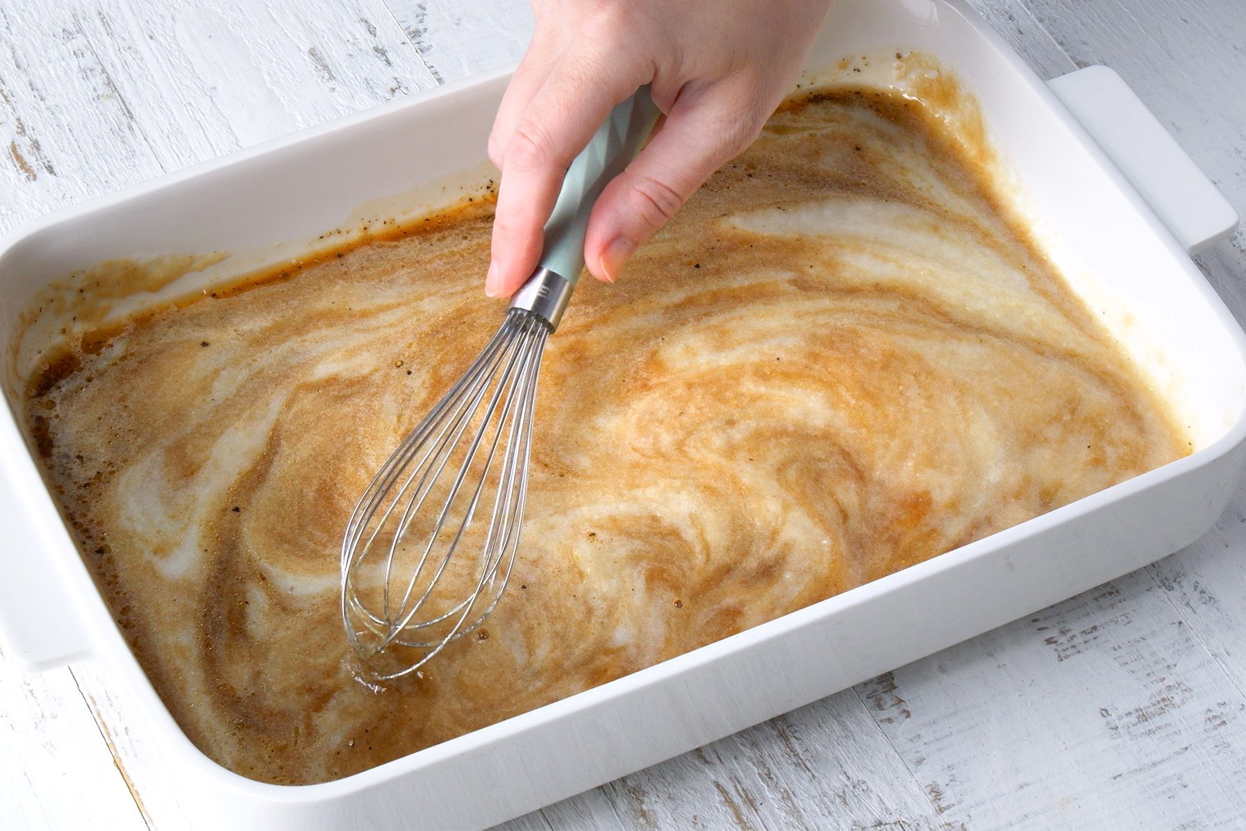 A hand using a whisk to stir a creamy, light brown mixture in a white rectangular baking dish placed on a distressed white wooden surface. 