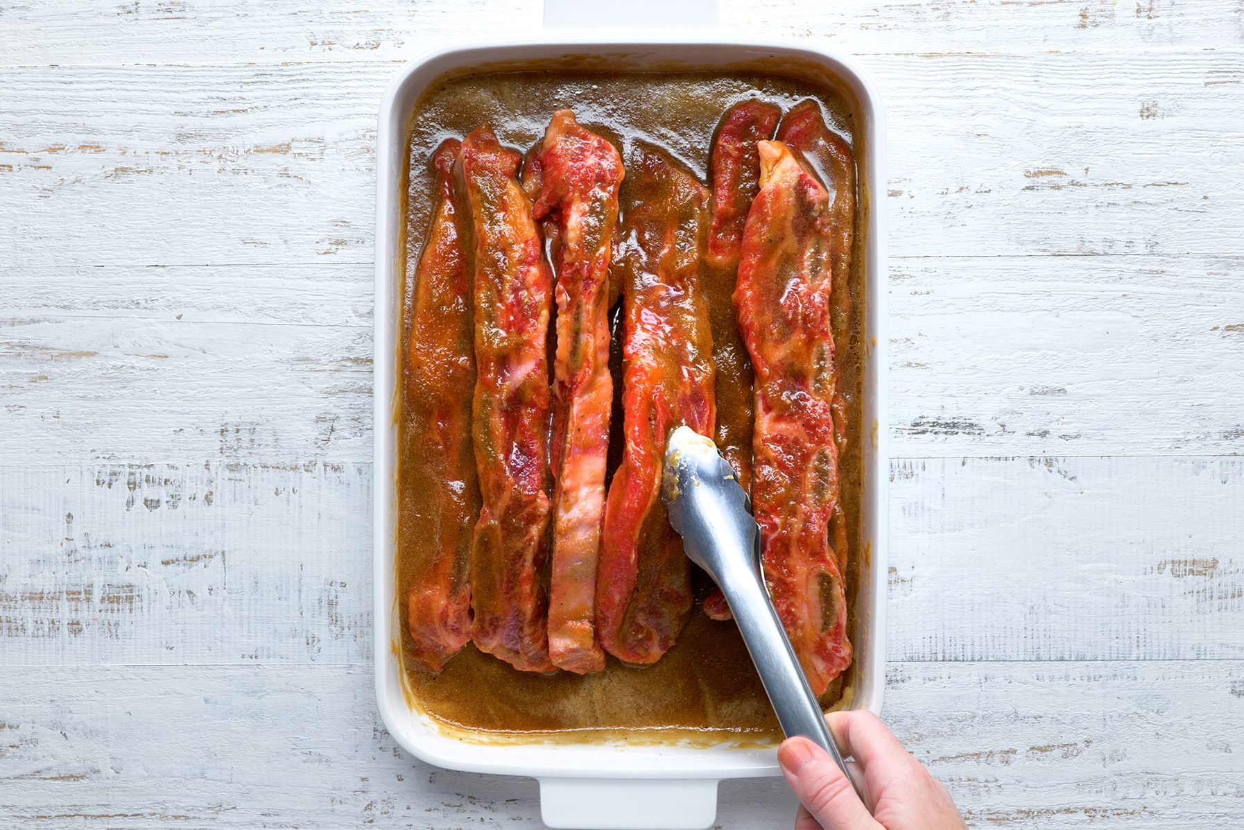 A hand uses tongs to place ribs in a white baking dish with a brown sauce. The dish is on a rustic white wooden surface. 