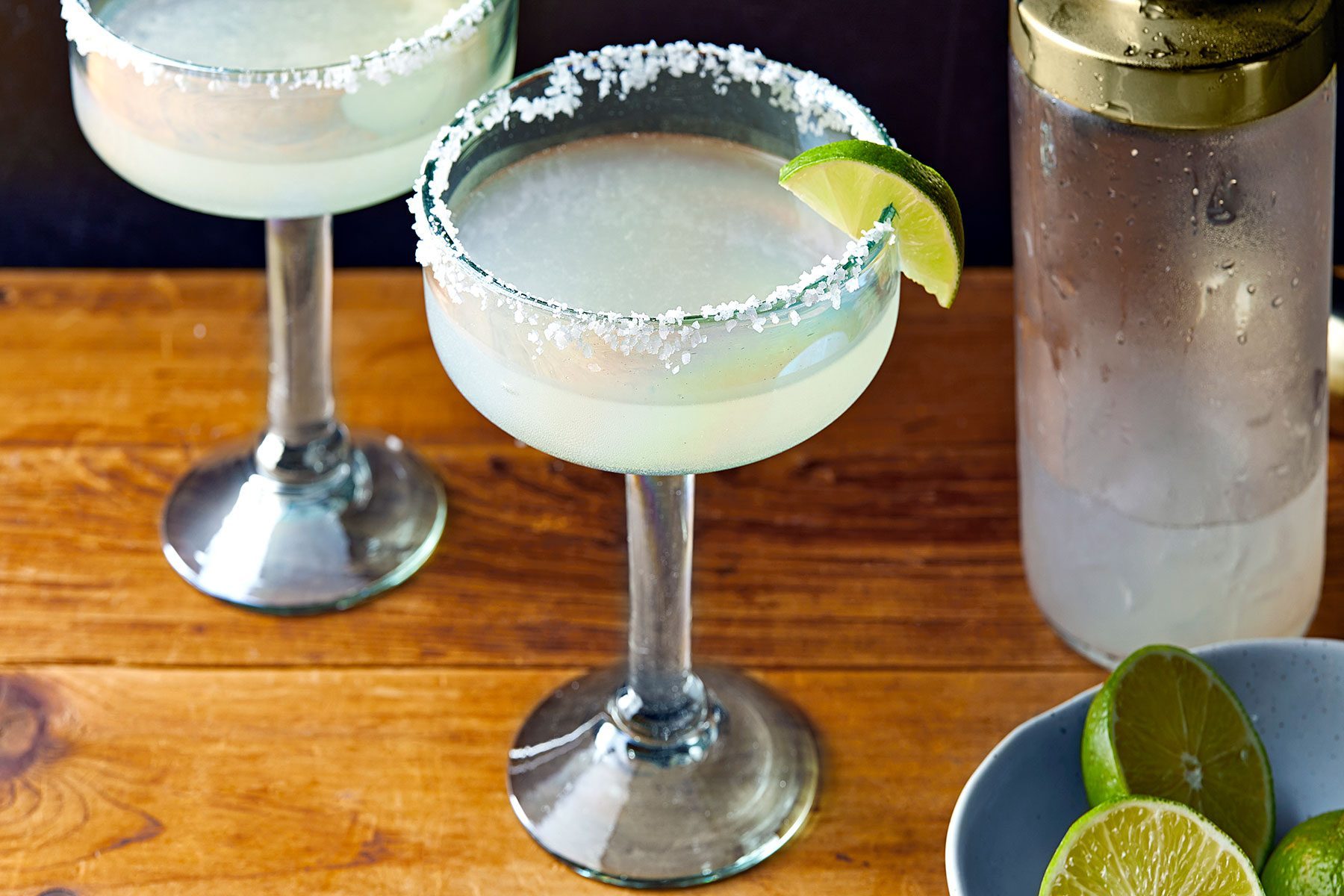 High angle view shot of Mezcal Margarita; in glasses; garnished with lime wedges; lime wedges bowl; shaker; wooden background