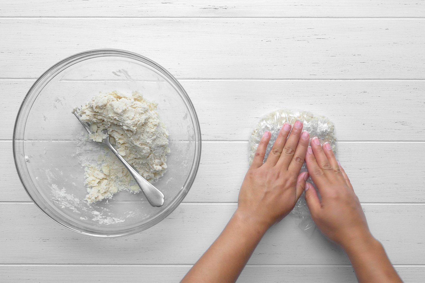 Hands pressing dough wrapped in plastic wrap on a white wooden surface. A clear bowl with a spoon and remaining dough sits to the left.