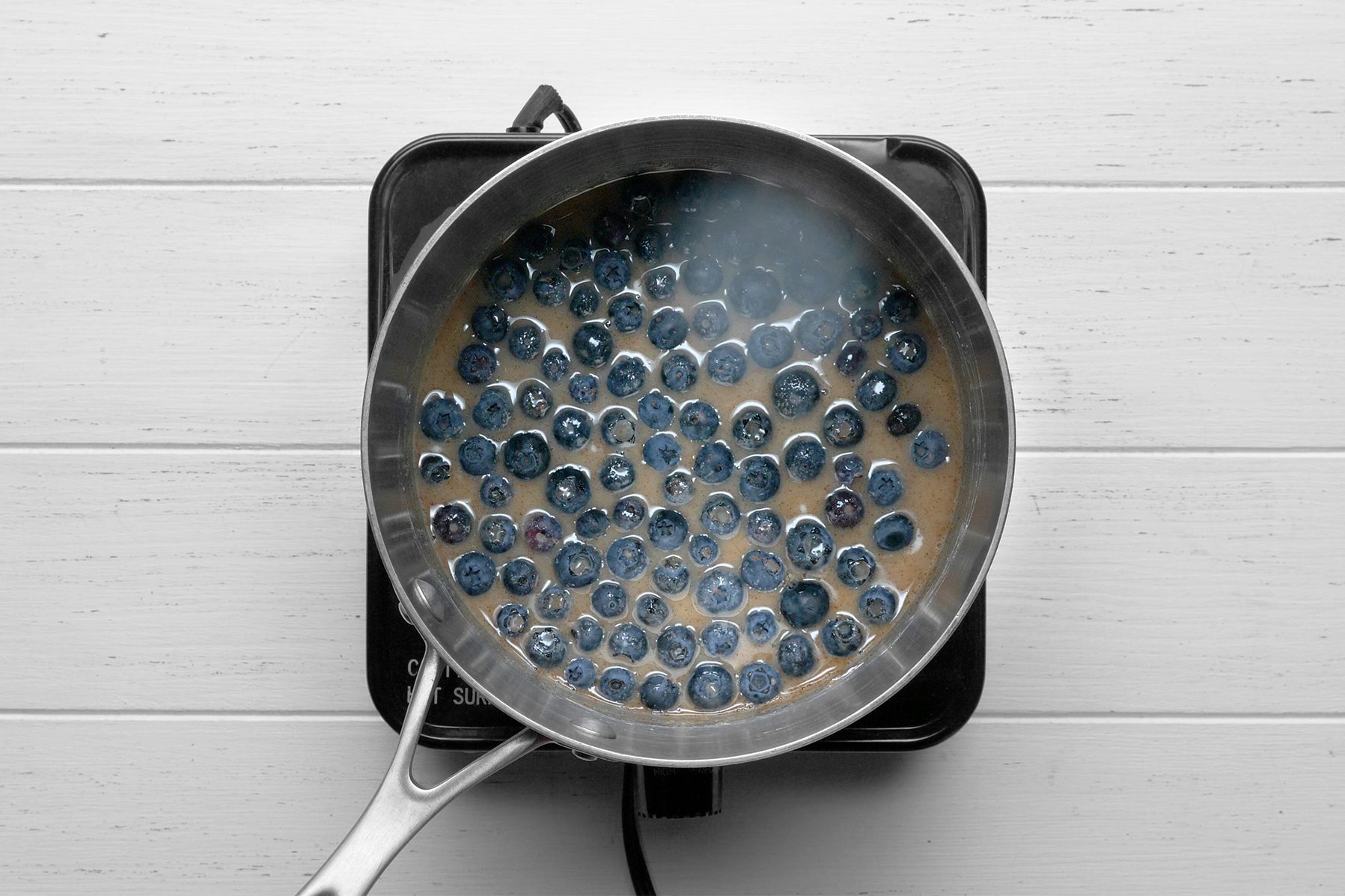 A stainless steel frying pan containing blueberries is on a small stovetop. 