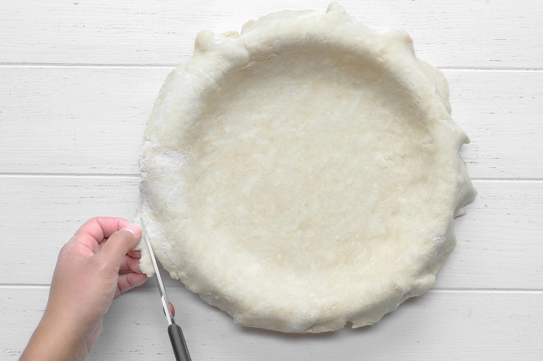 A person's hand is seen trimming the excess dough from the edge of an unbaked pie crust in a pie dish using a pair of scissors. 