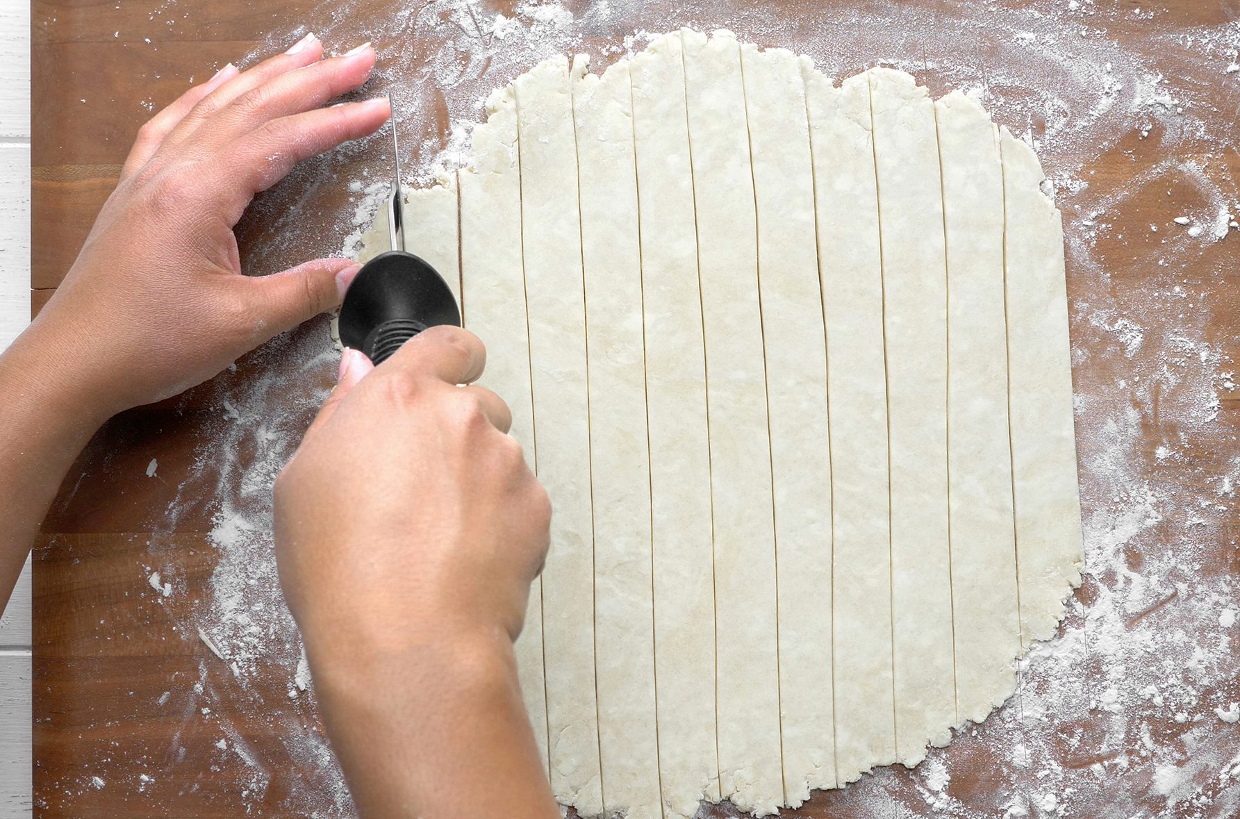 Two hands are cutting dough into strips on a floured wooden surface using a pastry wheel. 