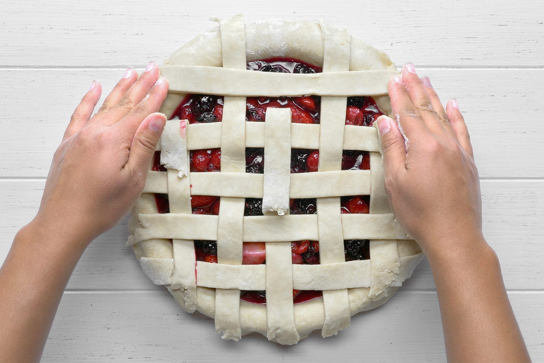 Two hands are weaving a lattice top crust over a fruit pie. The unbaked pie is filled with a mixed berry filling and is in the process of being prepared on a white wooden surface. 
