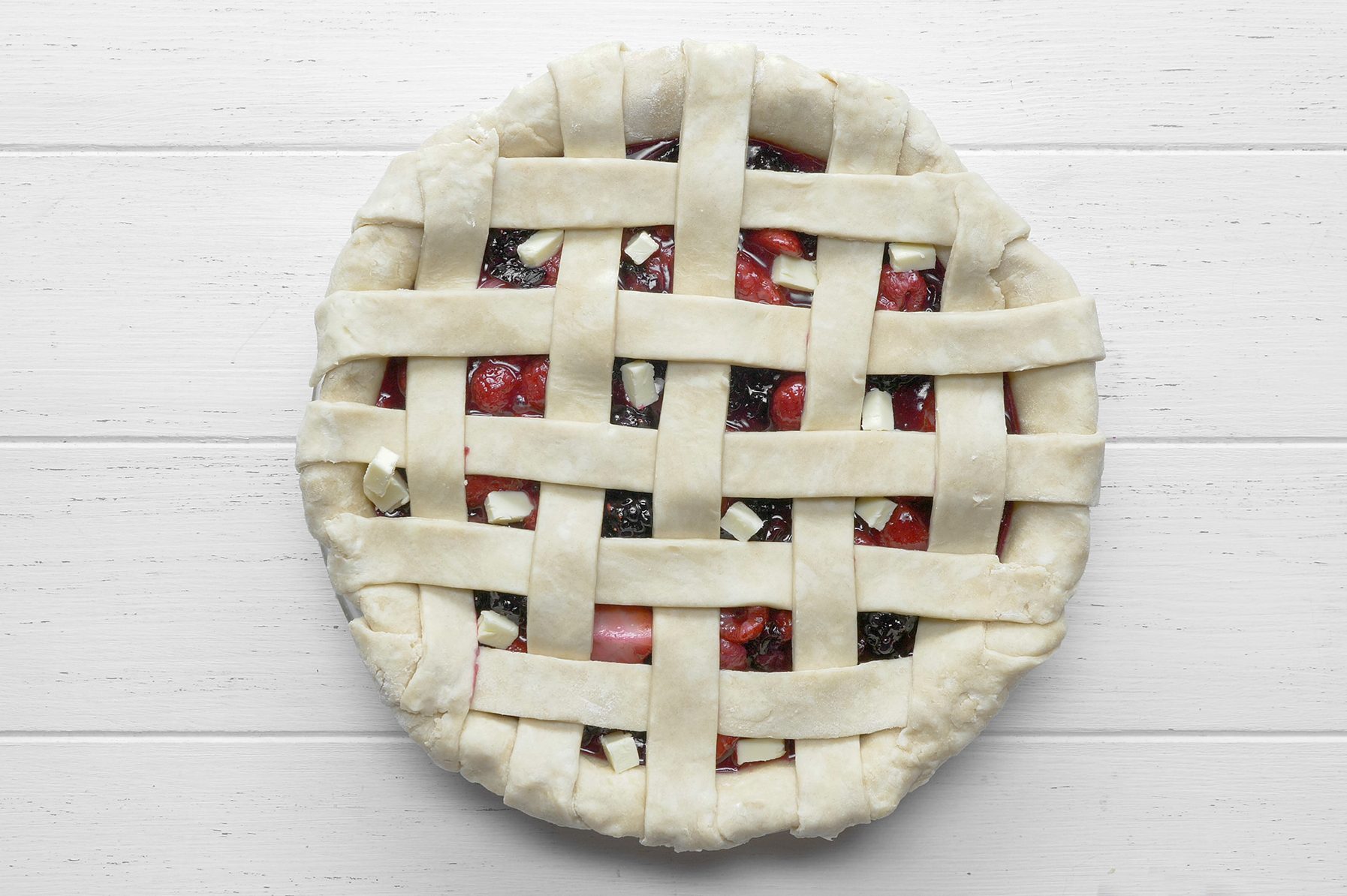 A top-down view of a lattice-topped cherry and berry pie on a white wooden surface. The golden-brown crust forms a woven pattern, revealing the red and dark purple fruits underneath. 
