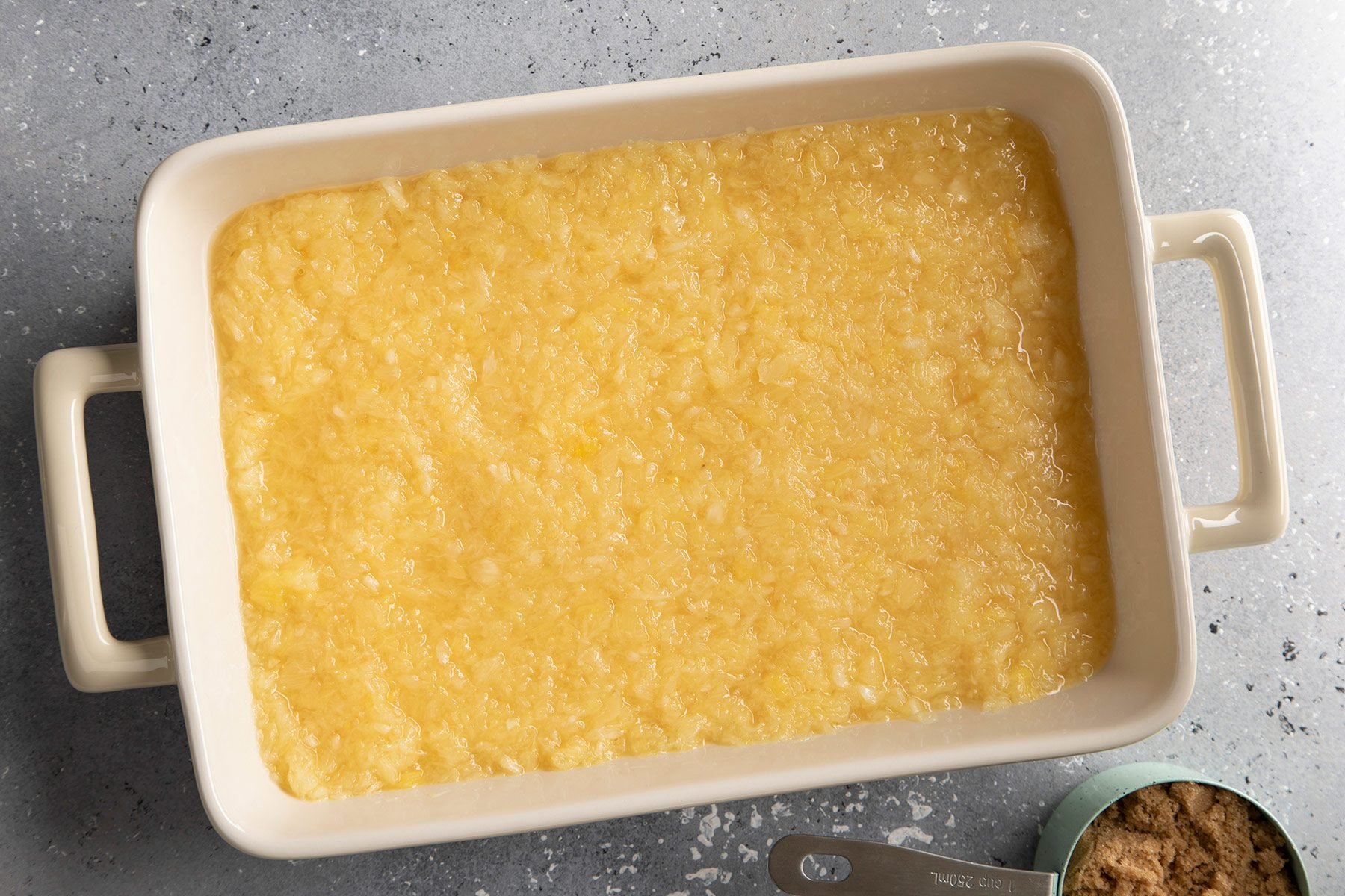 A rectangular baking dish filled with a yellow, textured mixture. The mixture appears to be evenly spread across the dish. A small, partially visible measuring spoon with a bit of a similar mixture is placed at the bottom right corner on a grey surface.