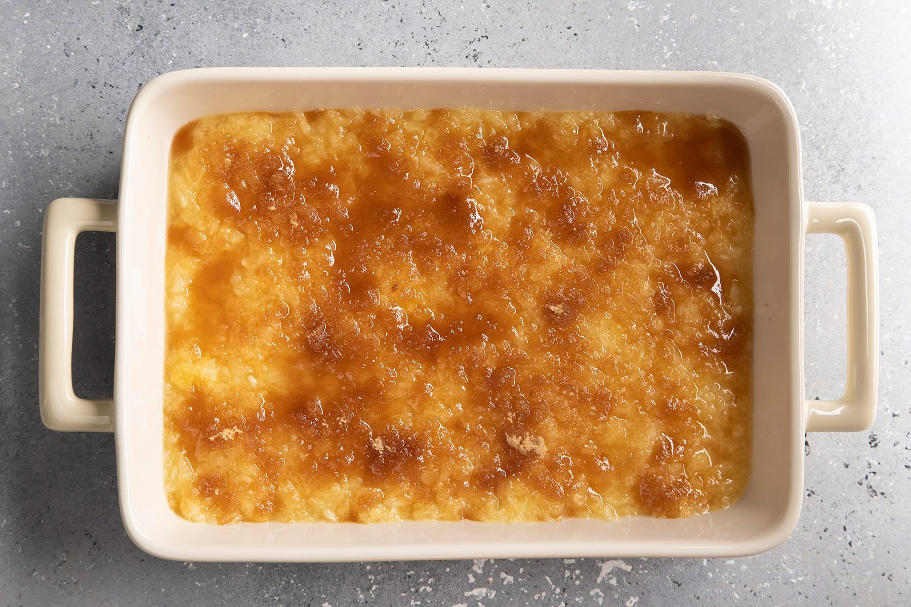 A rectangular baking dish filled with a golden-brown dessert, featuring a caramelized, slightly bubbly top layer. The dish appears to be placed on a lightly speckled grey surface.