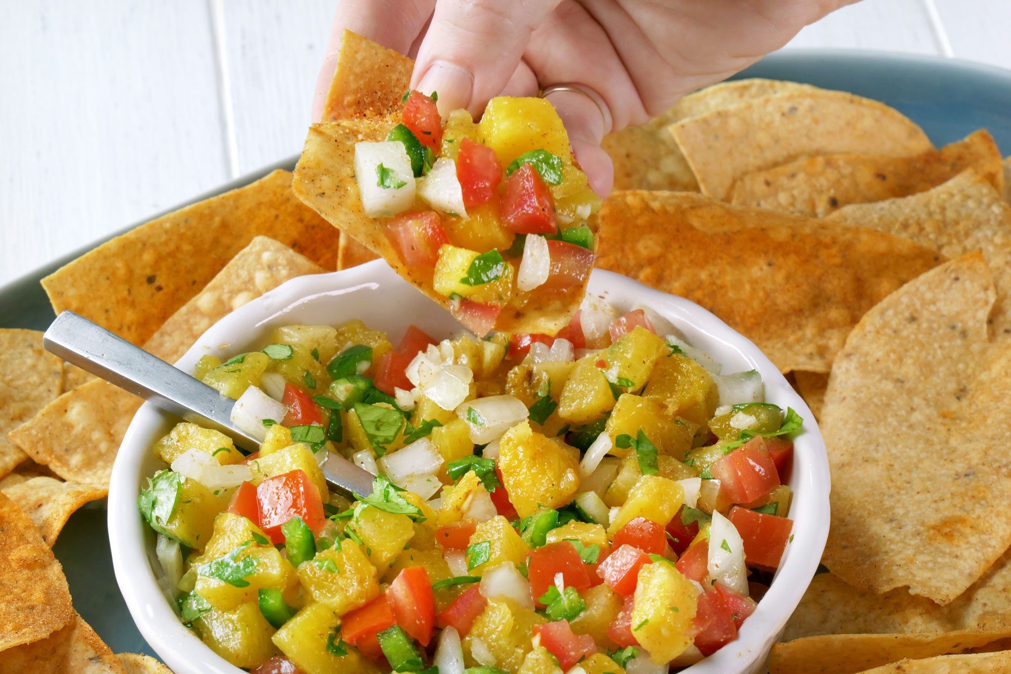 3/4th shot; white background; holding a tortilla chips with Pineapple Salsa over it; Pineapple Salsa in large white bowl served with tortilla chips in a blue plate;