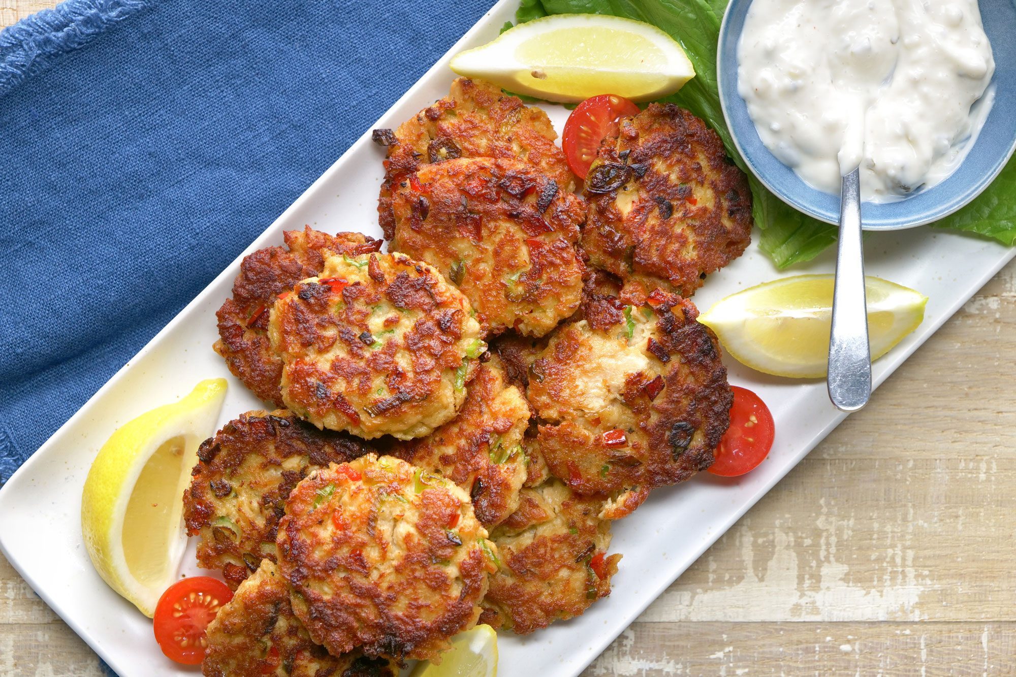 Overhead shot of Tasty Salmon Croquettes; serve on large platter; with tartar sauce and lemon wedges; spoon; cherry tomatoes; blue table cloth; wooden background;