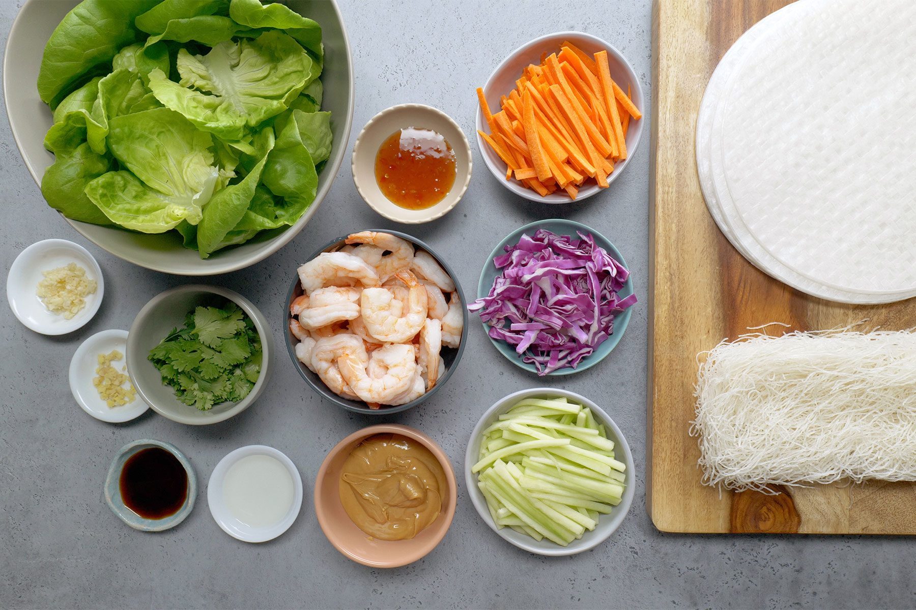 An assortment of ingredients for making Ingredients for Shrimp Spring Rolls is arranged on a surface. Items include lettuce, shrimp, sliced vegetables (carrots, purple cabbage, cucumber), sauces, herbs, and rice noodles, with rice paper sheets on a wooden board to the right.