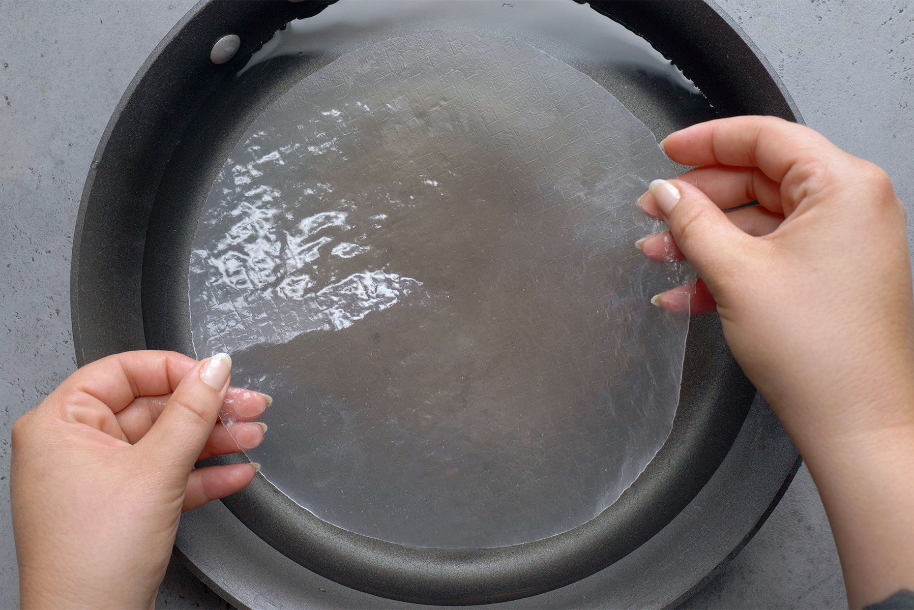 A person is holding a circular sheet of translucent rice paper with both hands above a shallow dish or pan filled with water. 