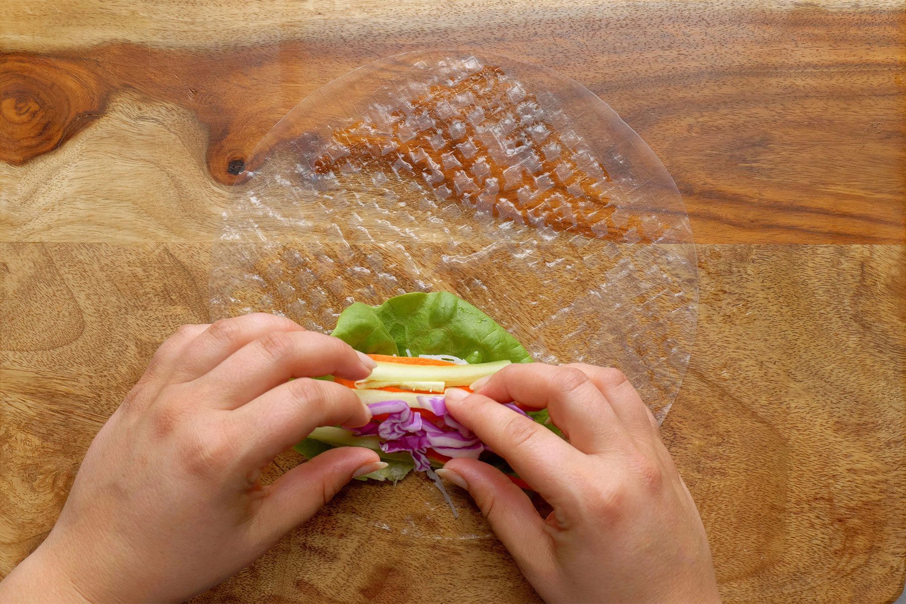 Two hands are shown folding a rice paper wrapper for a fresh spring roll. The wrapper contains a green leaf and slices of purple cabbage and orange vegetables. 