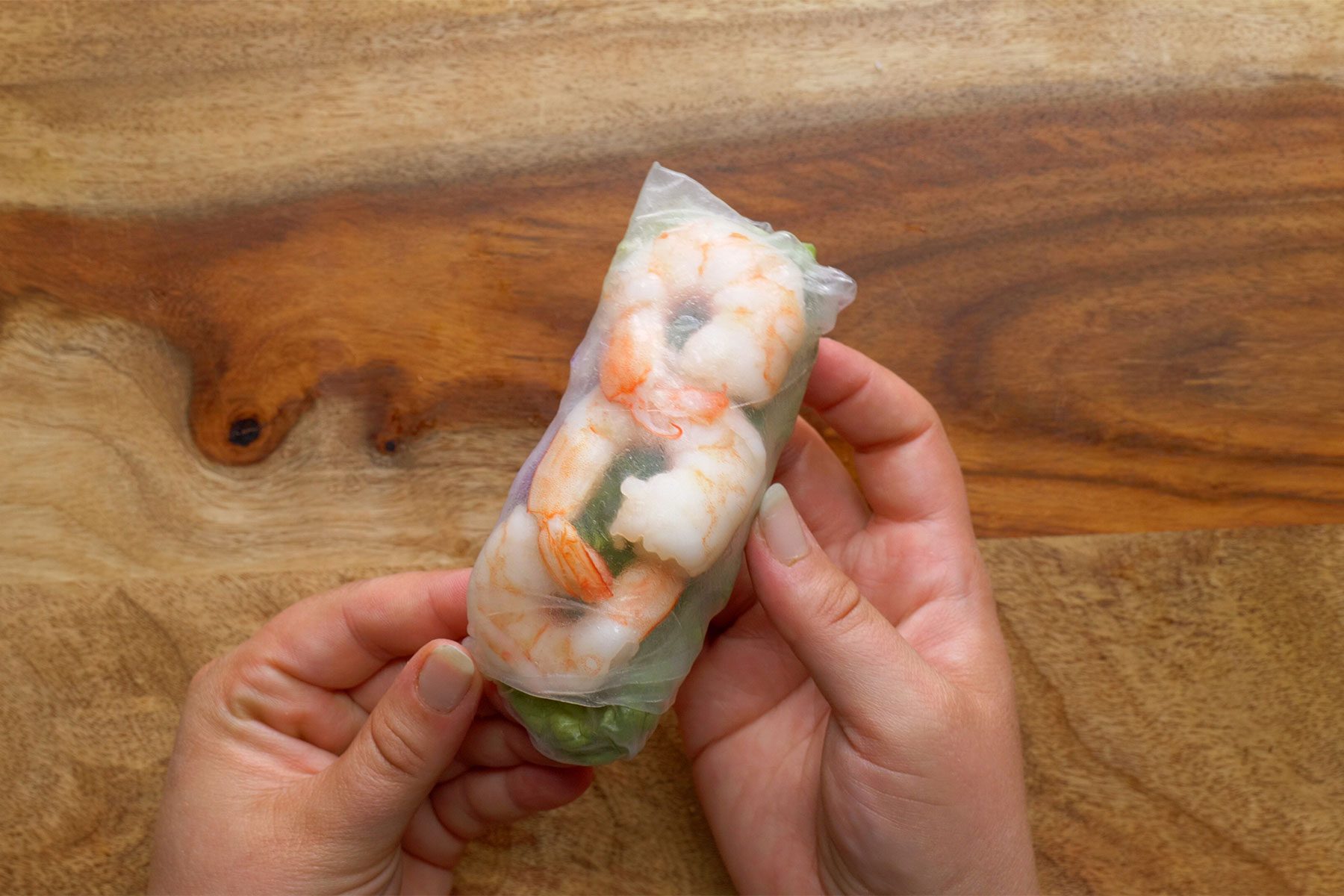 Two hands holding a fresh shrimp spring roll wrapped in translucent rice paper, showcasing pieces of shrimp, lettuce, and other vegetables inside. The background is a wooden surface.