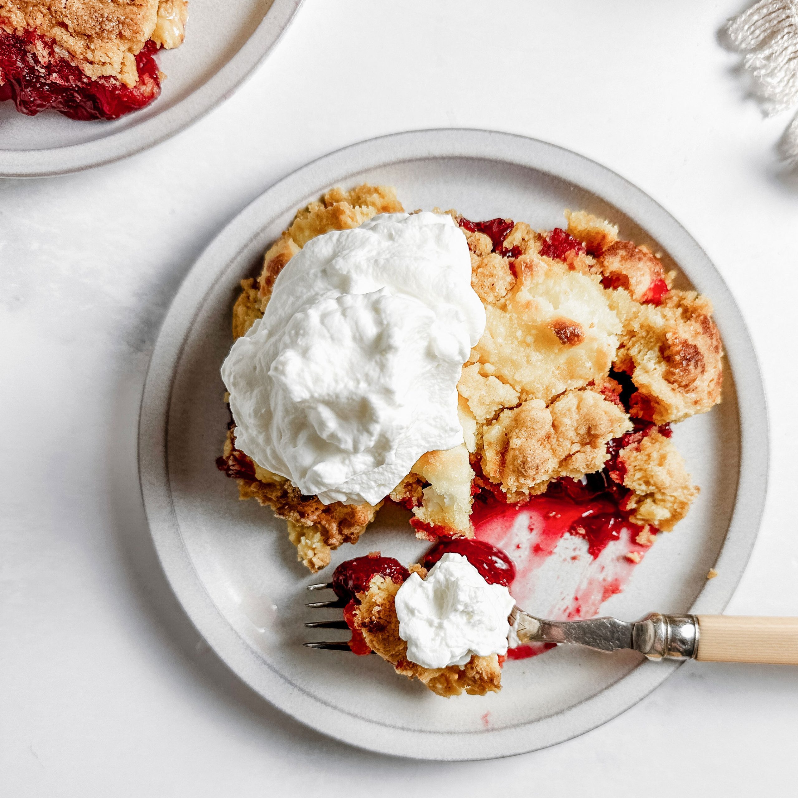 Taste of Home Strawberry Cheesecake Dump Cake topped with whipped cream on ceramic plates next to a cloth napkin.