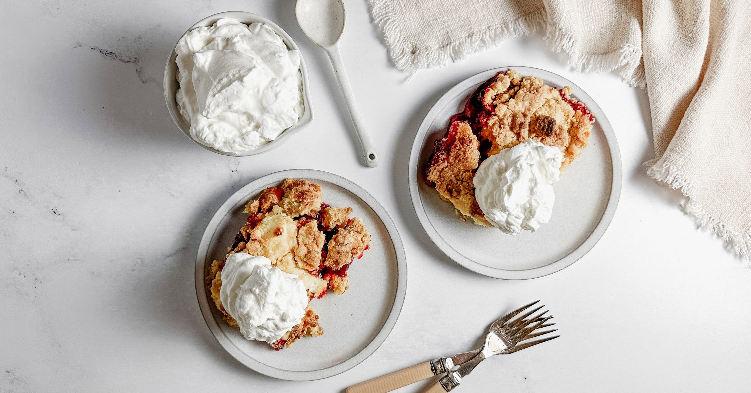 Taste of Home Strawberry Cheesecake Dump Cake topped with whipped cream on ceramic plates next to a bowl of whipped cream and a cloth napkin.