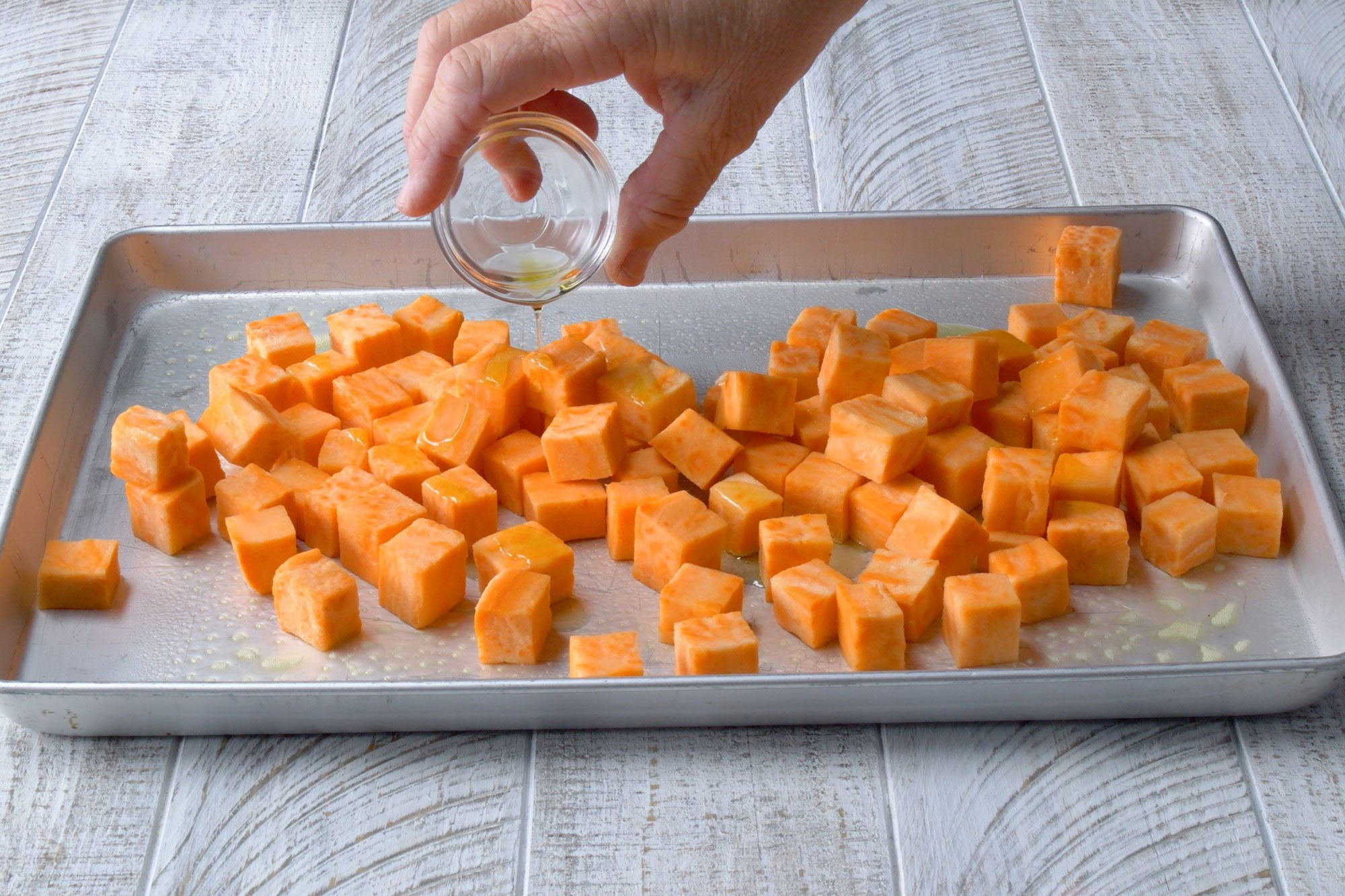 wide shot of cubed potatoes on a baking tray, sprinkle oil; white wooden background;