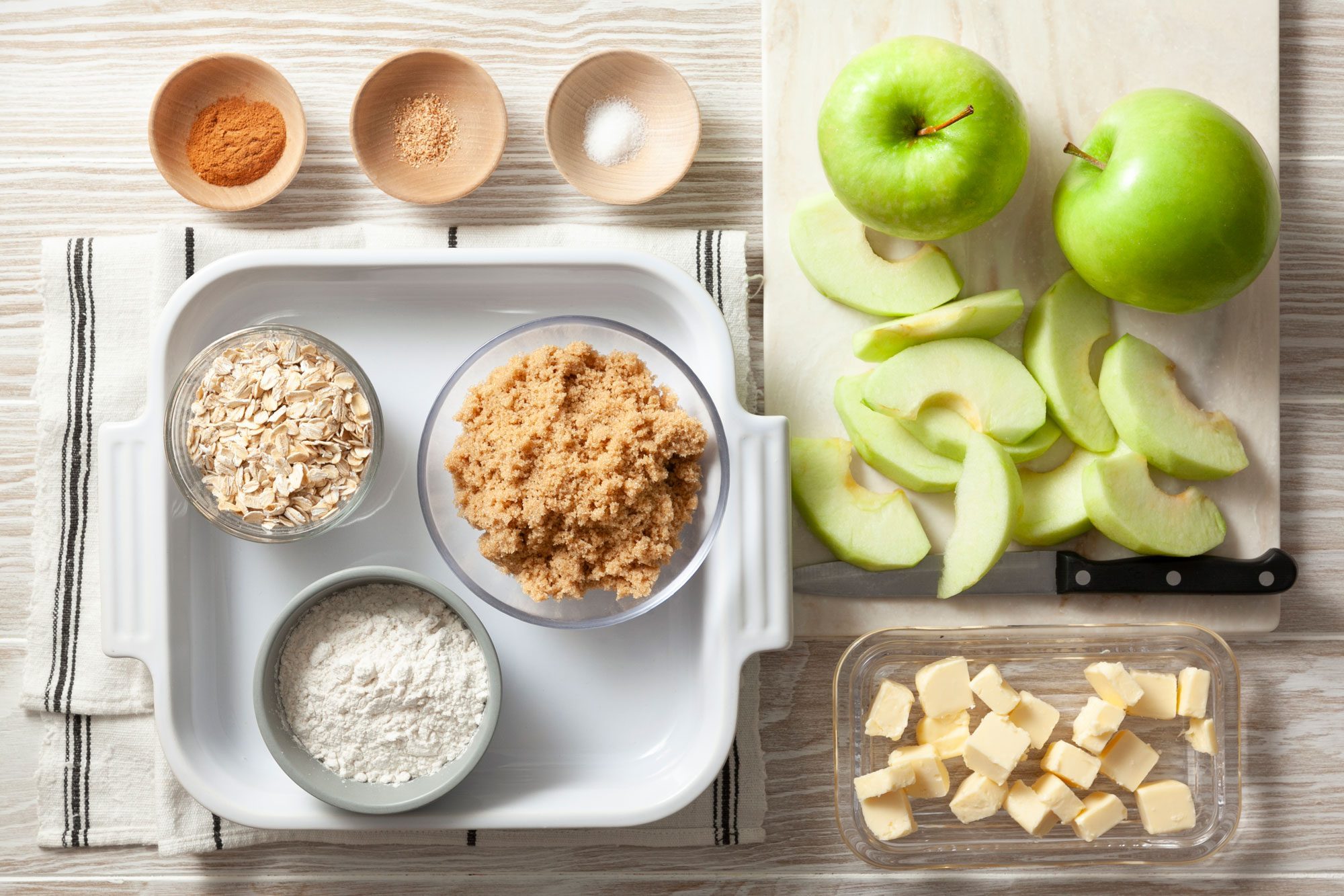 overhead shot; wooden background; Vegan Apple Crisp ingredients placed over background with white kitchen towel and knife;