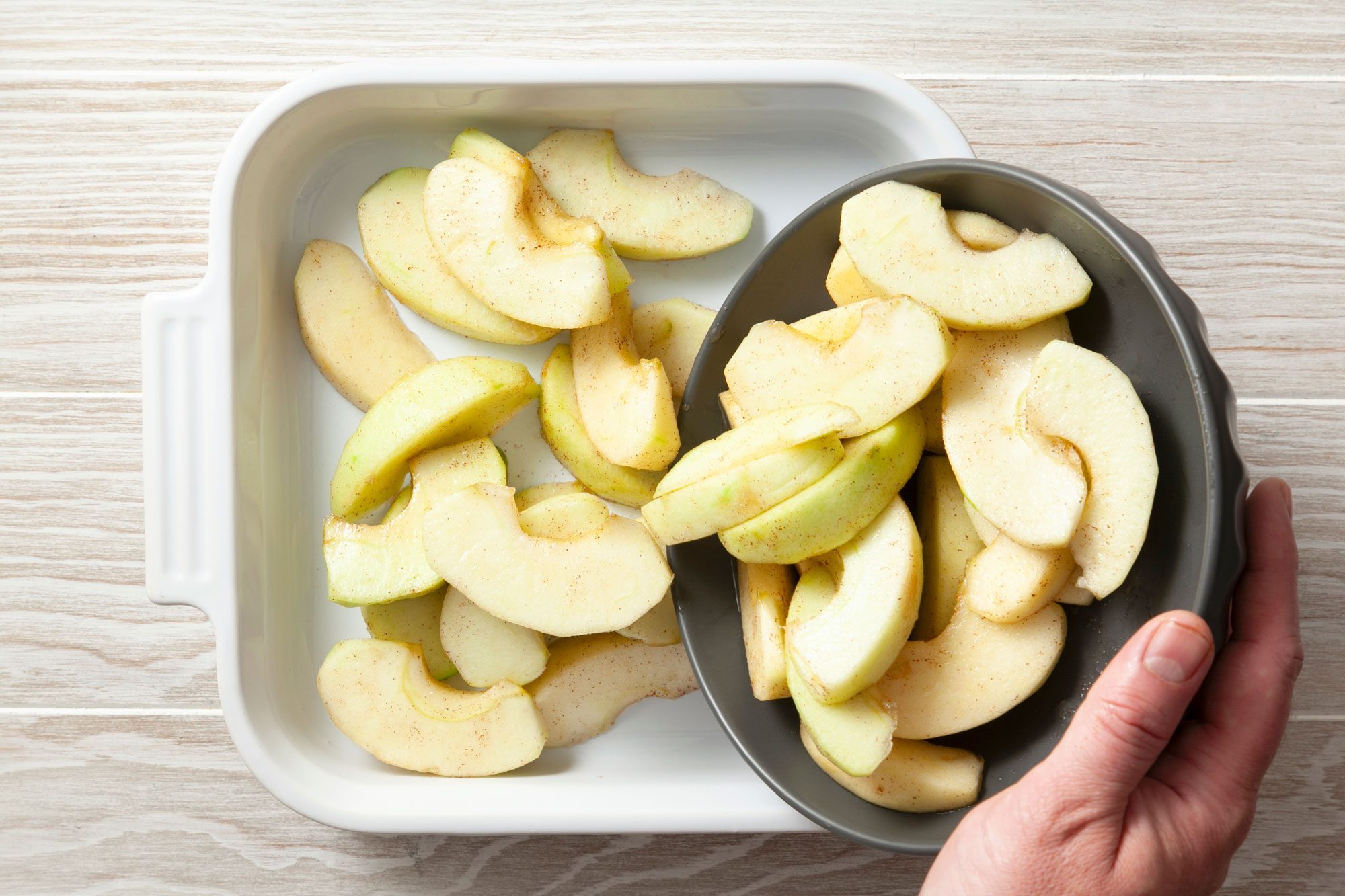 overhead shot; wooden background; transferring apple slices from a bowl to a square baking dish;