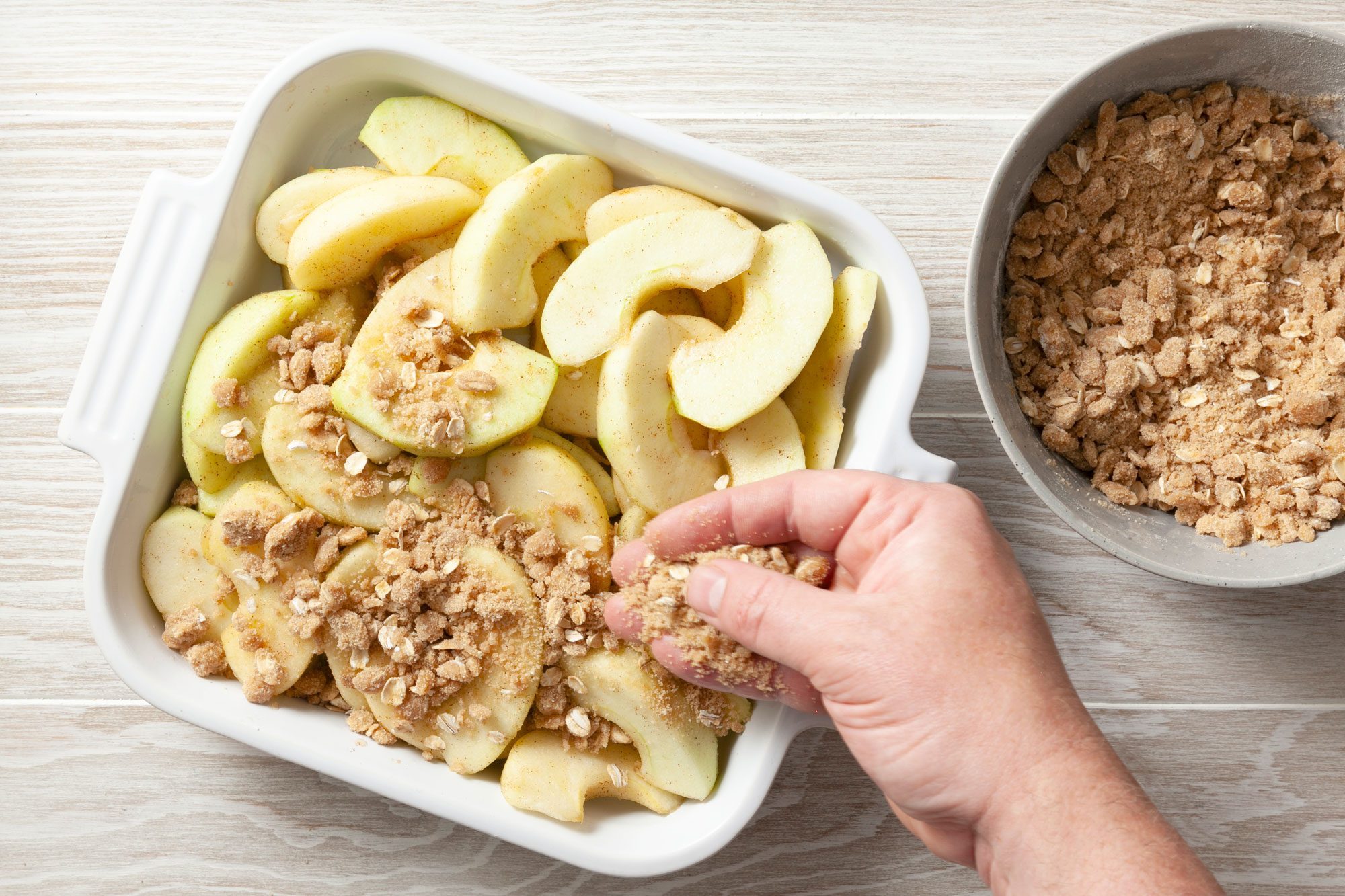 overhead shot; wooden background; sprinkling the oats mixture over apple slices in square baking dish;